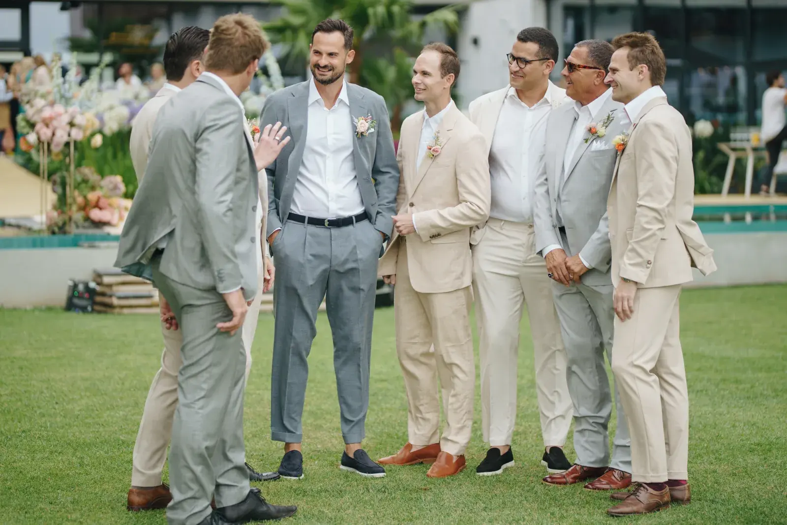 Groomsmen gathered on the ceremony lawn in pale linen, pool and florals behind them