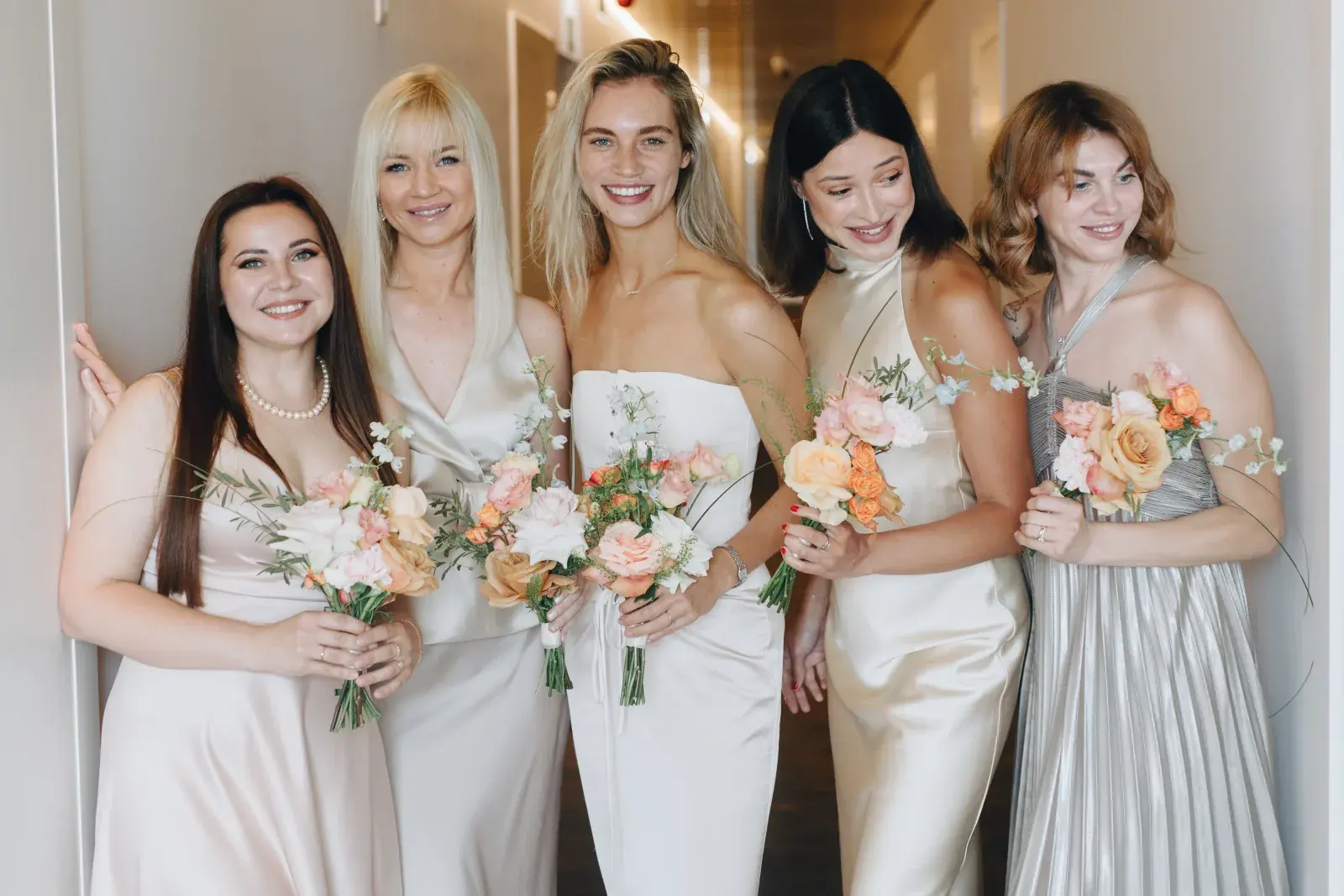 Bridesmaids in champagne satin holding peach bouquets in the corridor before the first look