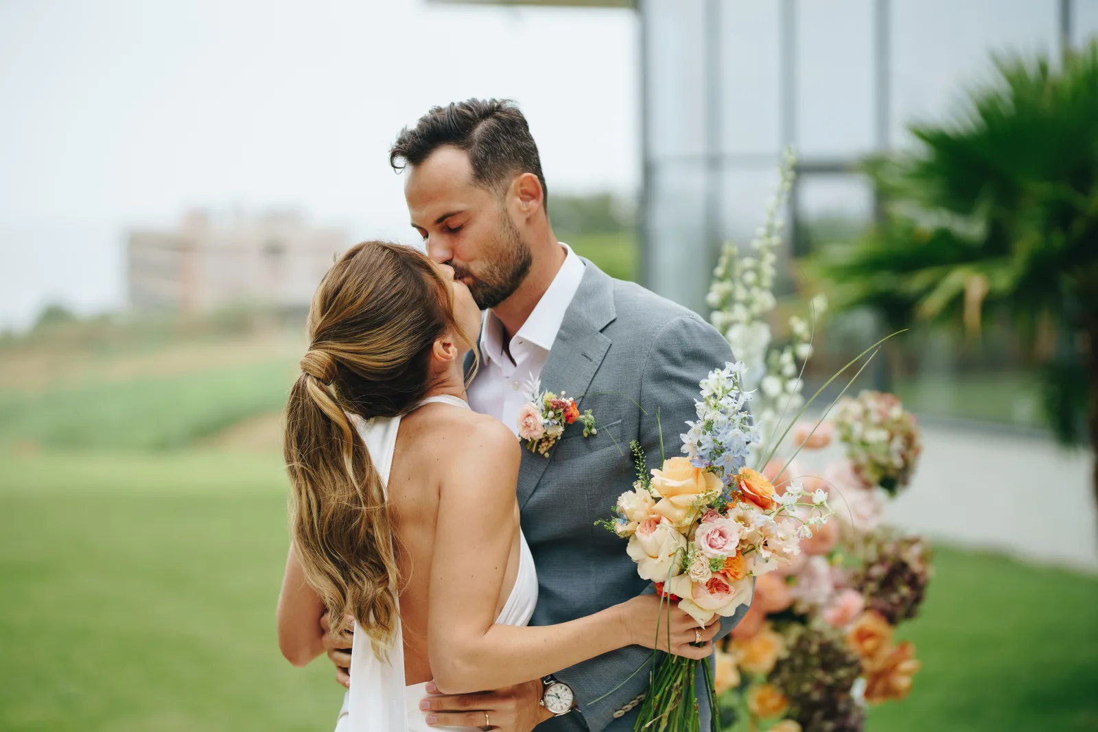 Couple kissing on the lawn with a pastel bouquet between them, glass pavilion and palms behind
