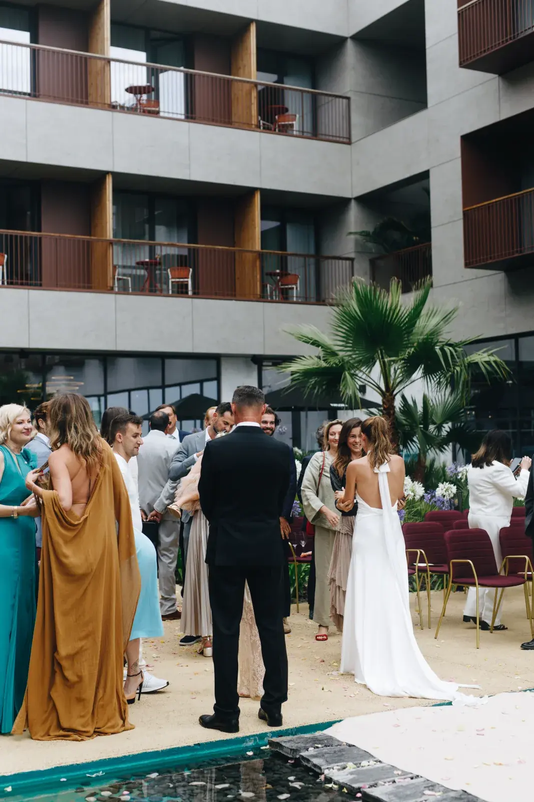 Guests mingling by the dark reflecting pool after a courtyard ceremony, petals on the water