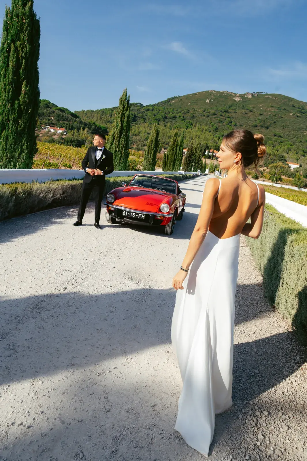 Bride in a backless gown on the estate drive, the groom waiting by a red vintage roadster among cypresses