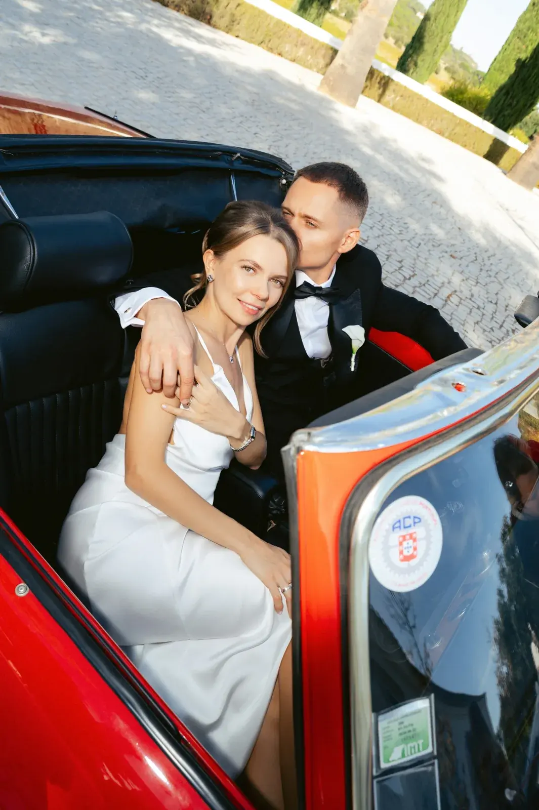 Bride and groom settled in the red vintage cabriolet on a cobbled drive, cypresses beyond