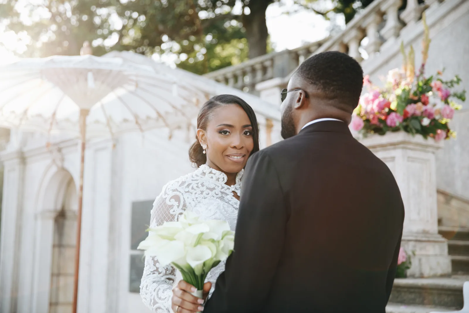Bride with calla lilies glancing back over the groom's shoulder, parasol and stone balustrade behind