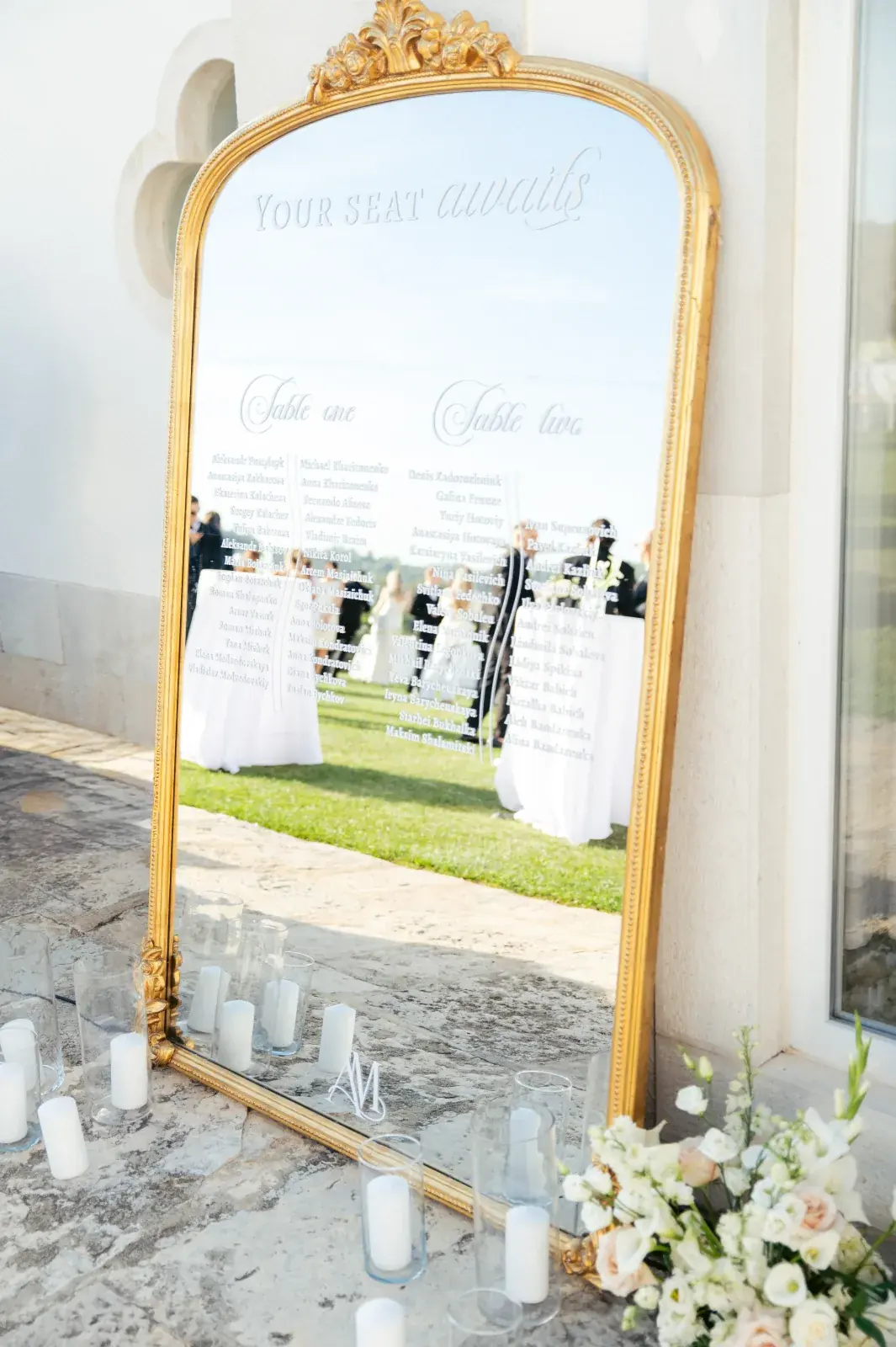 Gilt-framed mirror seating plan reading Your Seat Awaits, pillar candles and white roses at its base
