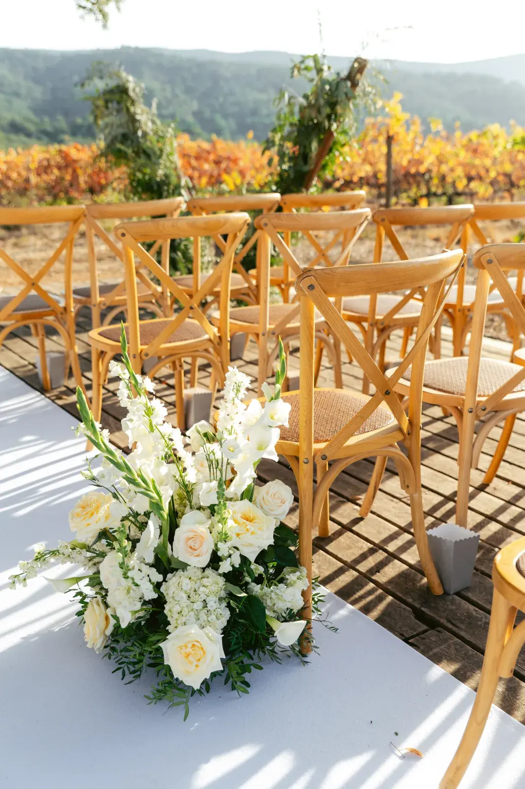 Aisle arrangement of cream roses and stocks beside wooden crossback chairs, autumn vines beyond