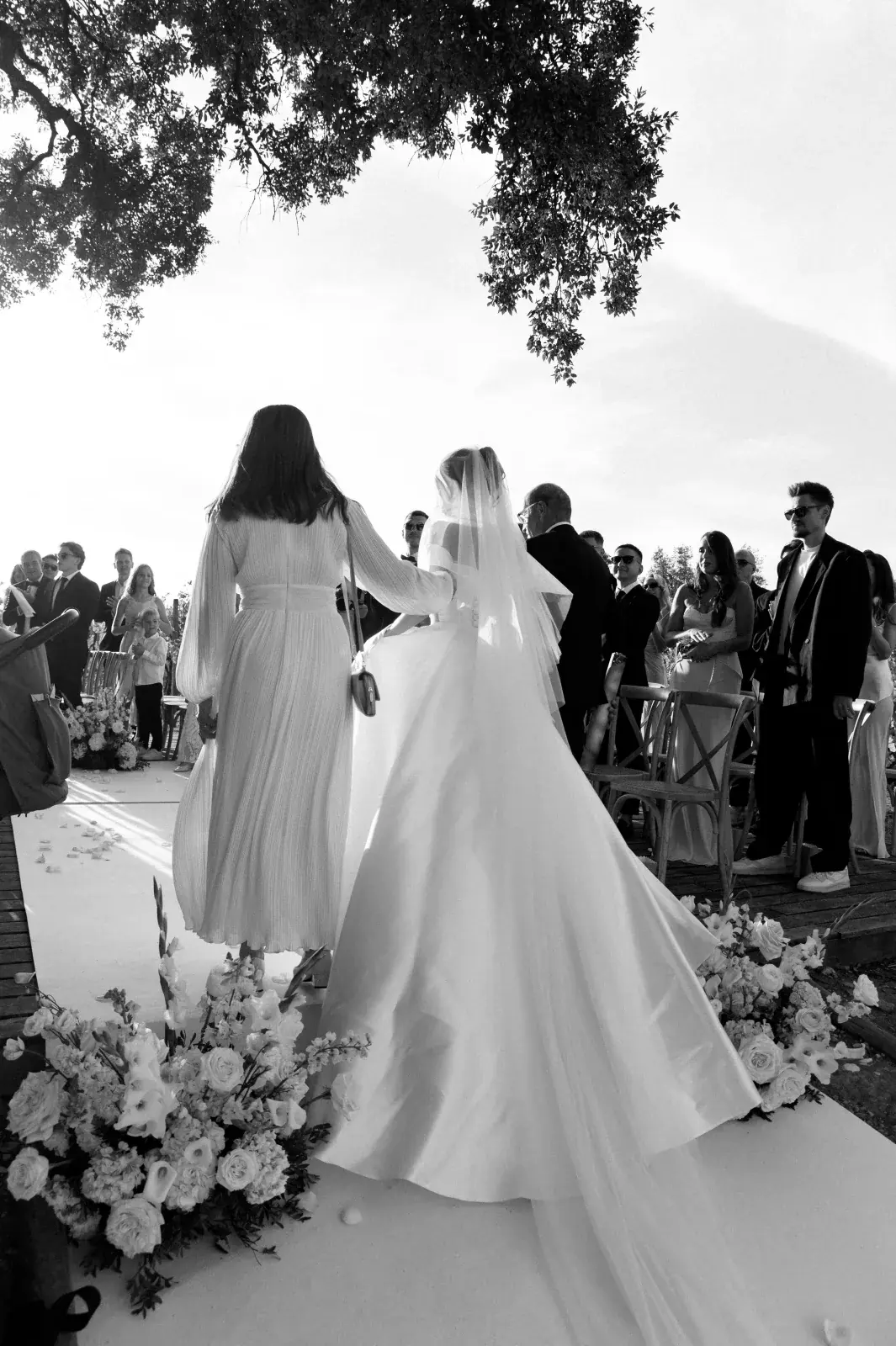 Bride escorted down a petal-lined aisle between standing guests at an open-air ceremony, in black and white