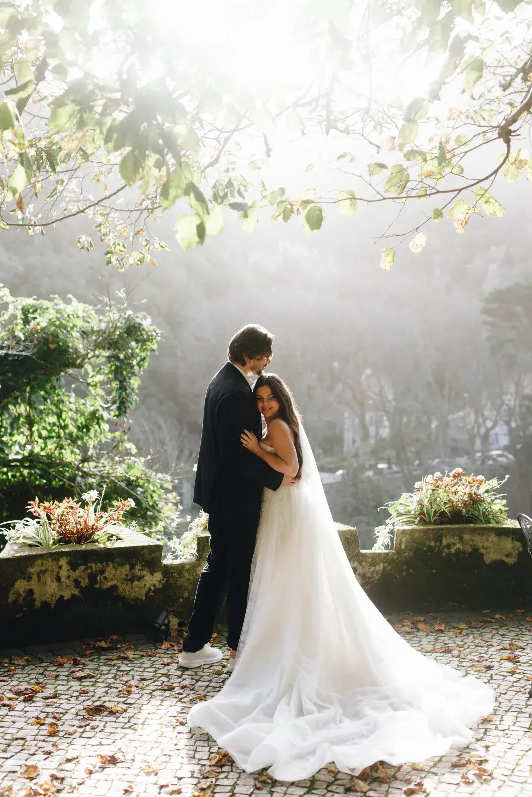 Couple embracing on a mossy stone terrace above town rooftops, morning mist softening the hillside light