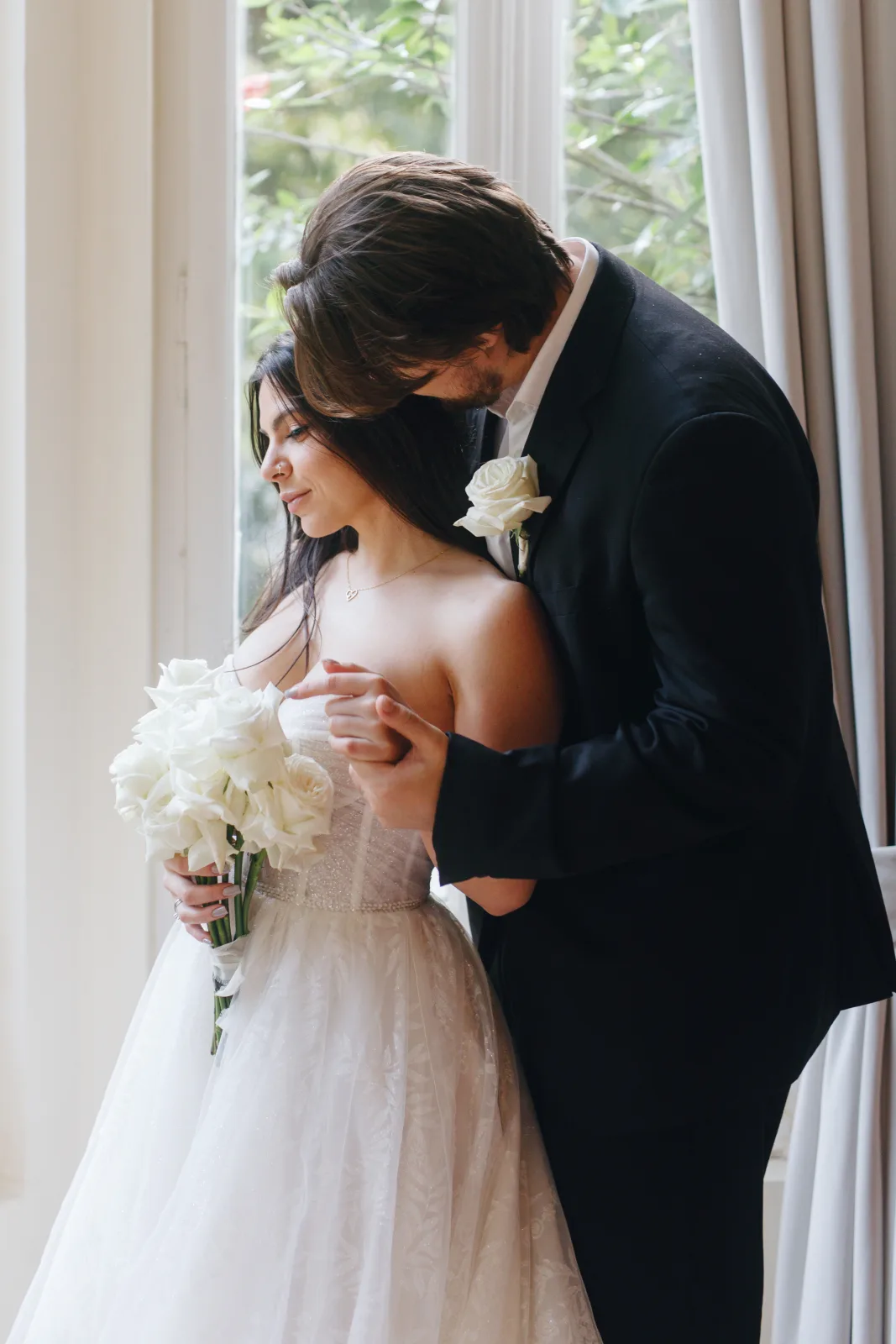 Groom holding the bride from behind at a tall window, white roses in her hand and soft interior light