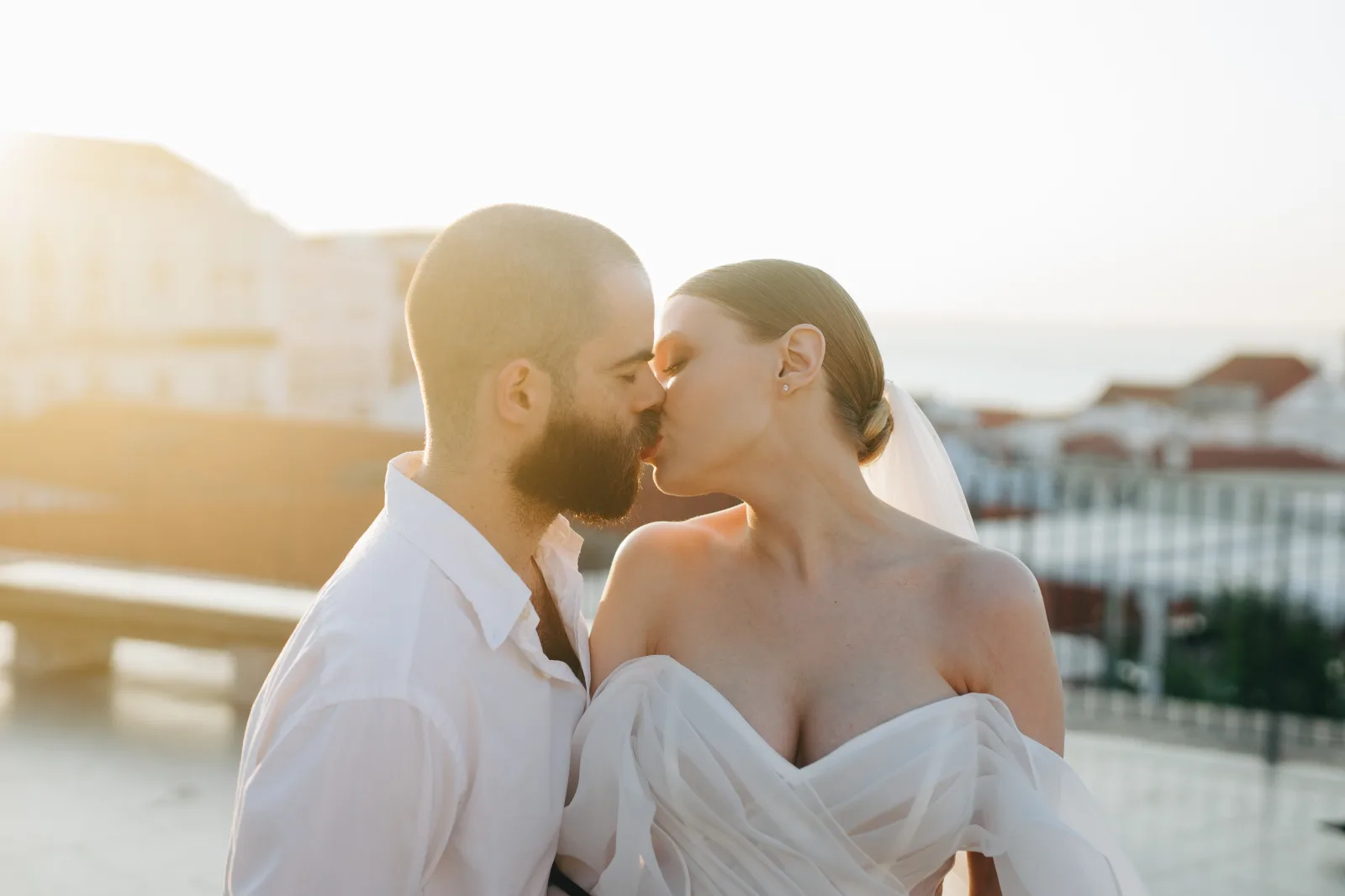 Rooftop kiss at golden hour above whitewashed town roofs, the sea pale behind