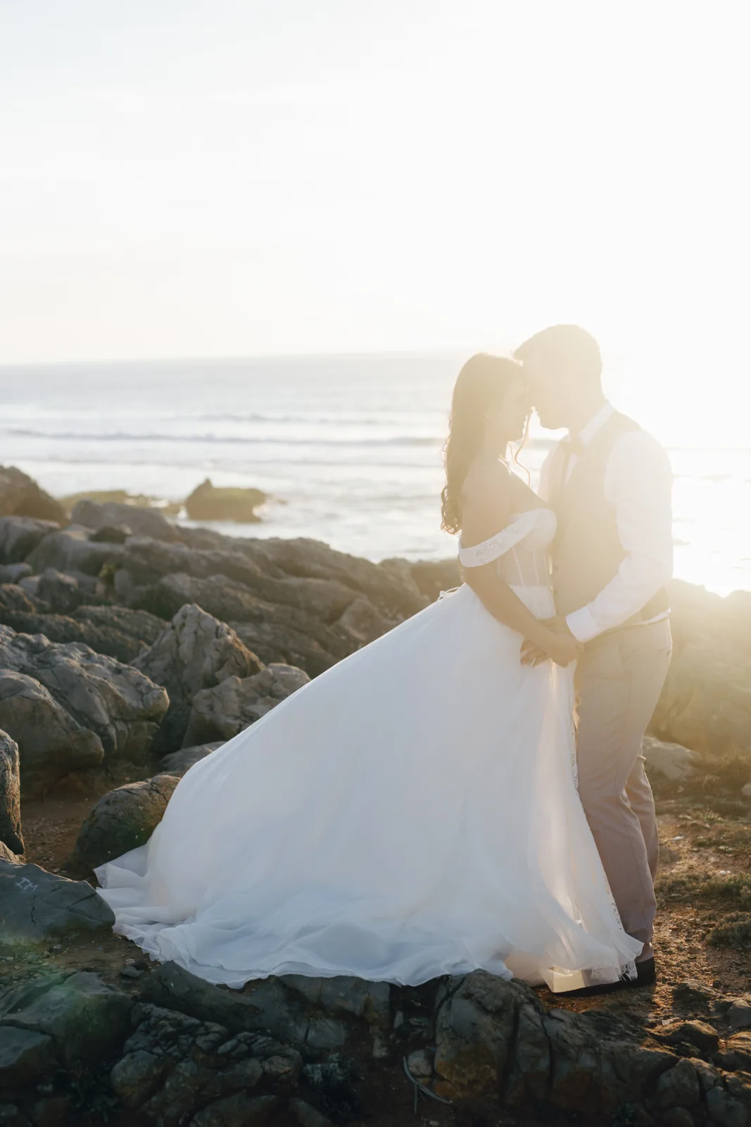 Bride and groom kissing on Atlantic rocks at sunset, her skirt spread across the stone