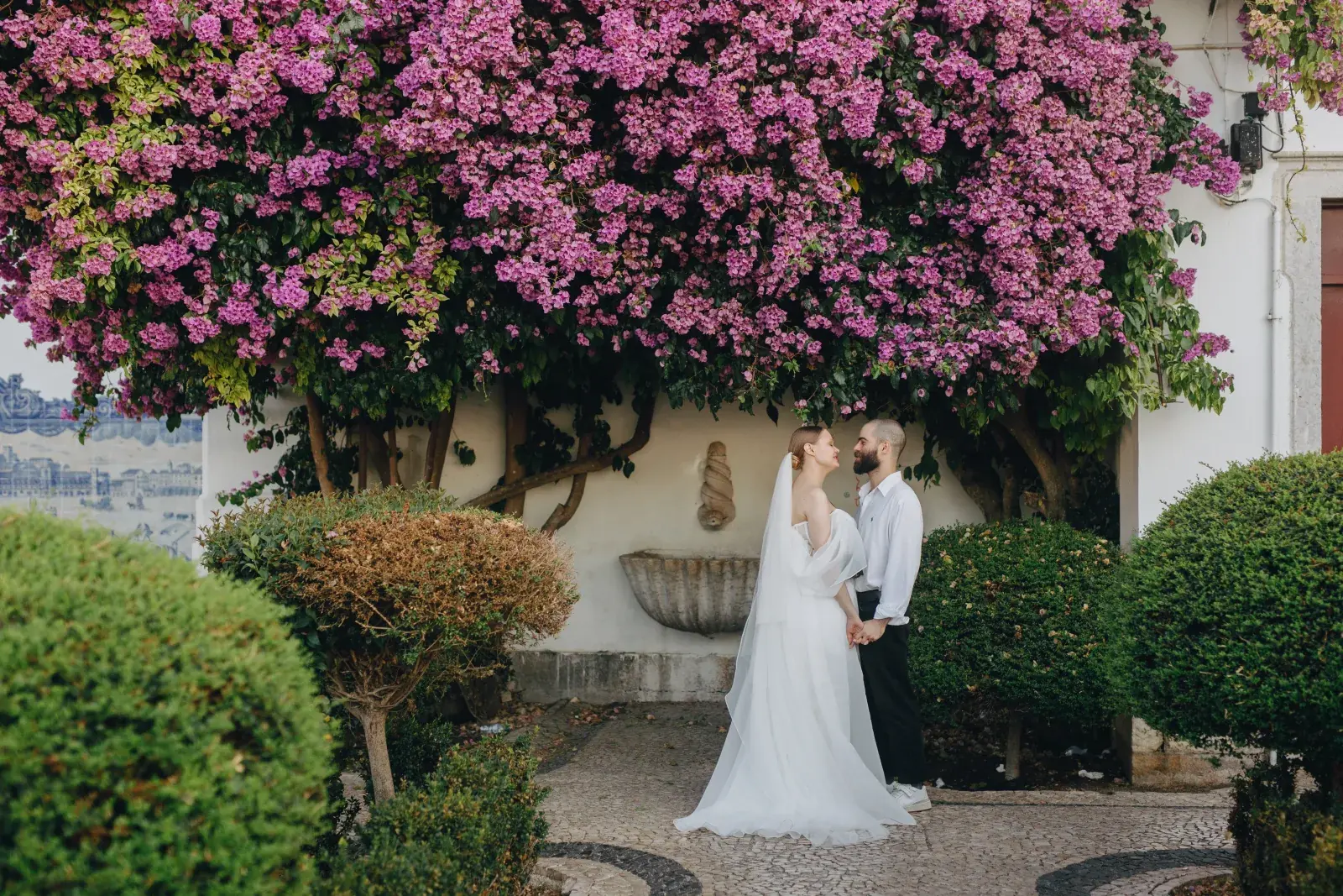 Couple hand in hand beneath pink bougainvillea spilling over a whitewashed courtyard wall