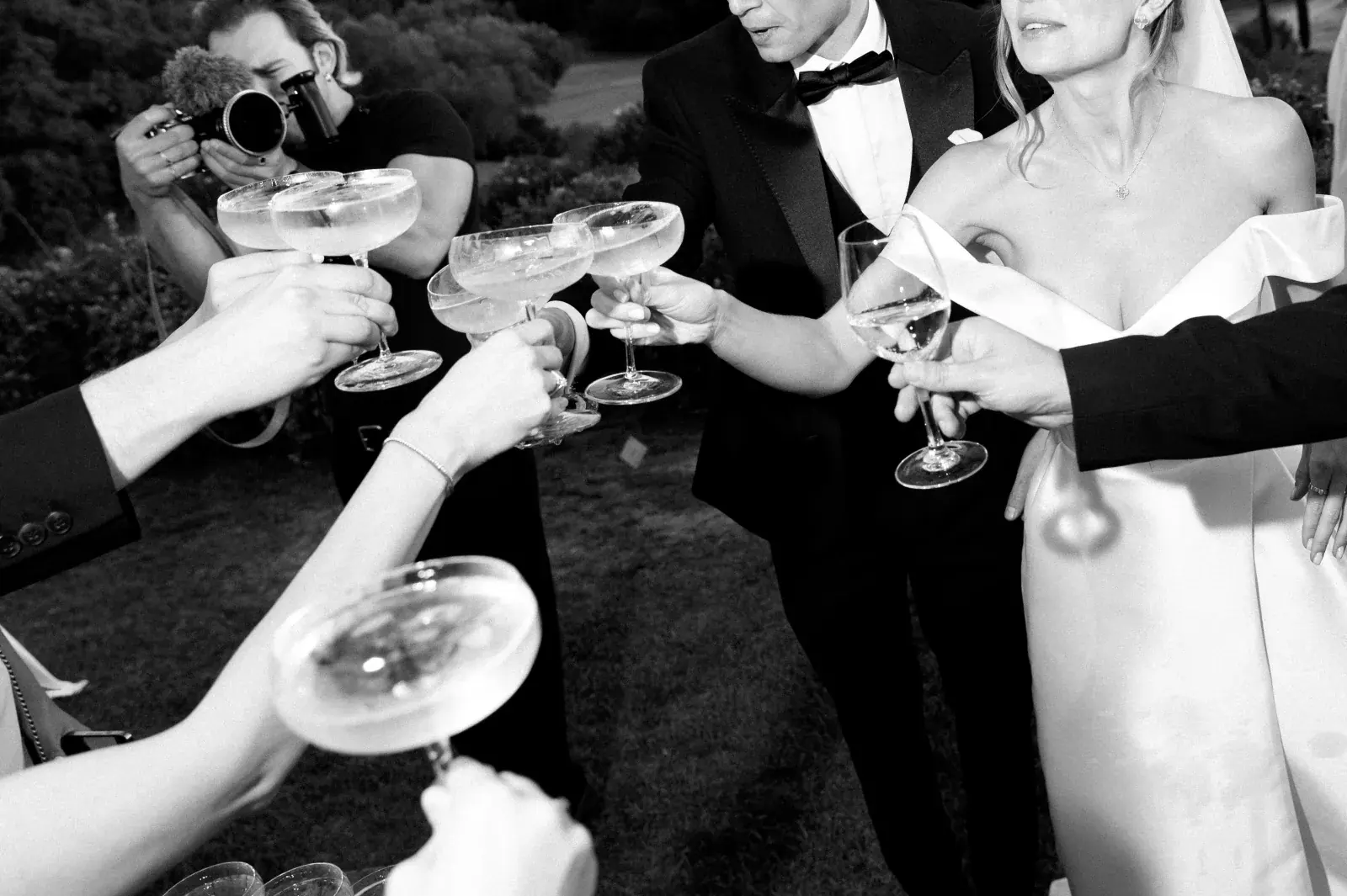 Champagne coupes raised in a toast on the lawn, the videographer leaning in, black and white
