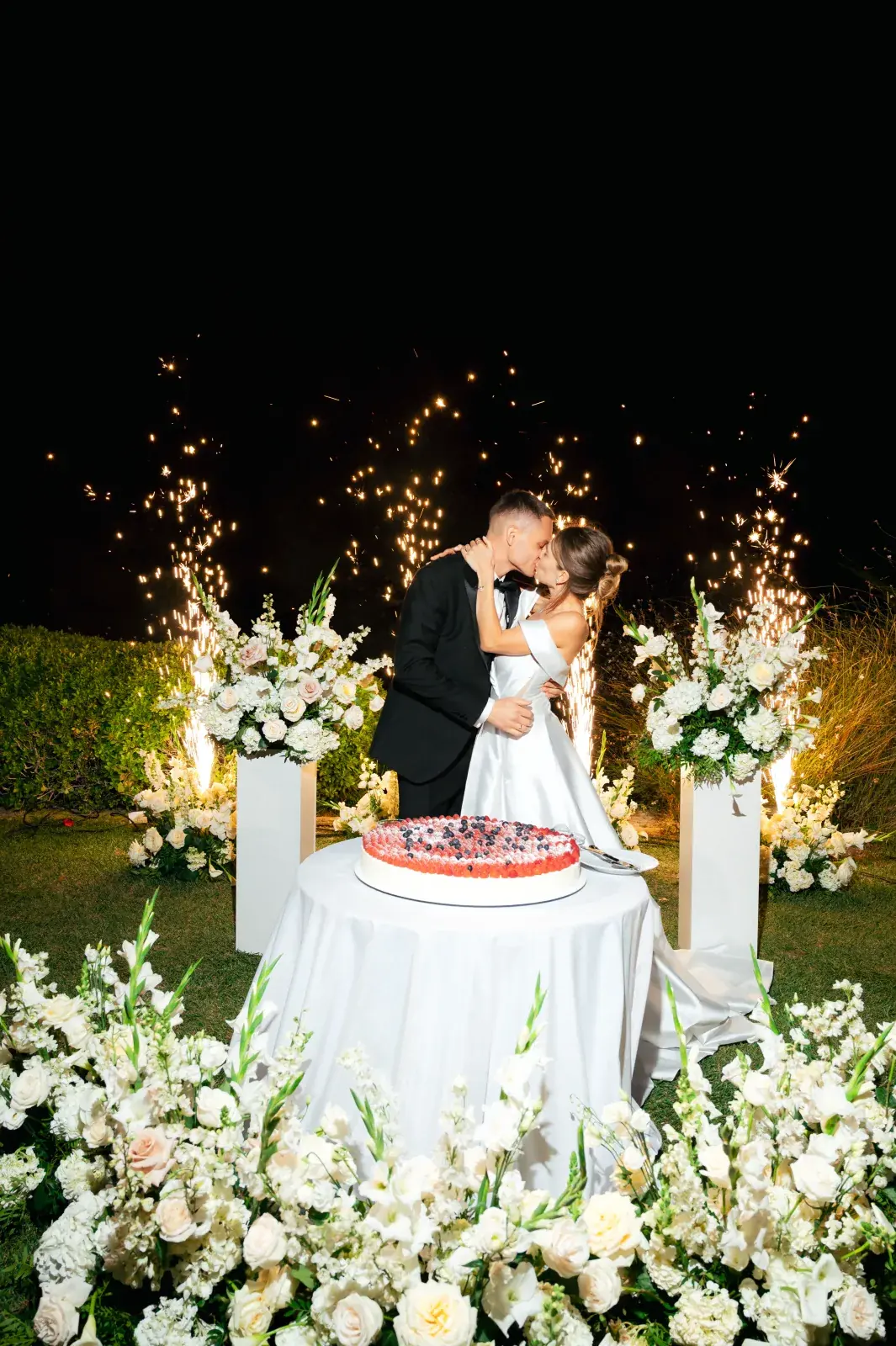 Newlyweds kissing behind the cake table as cold-spark fountains light the night lawn