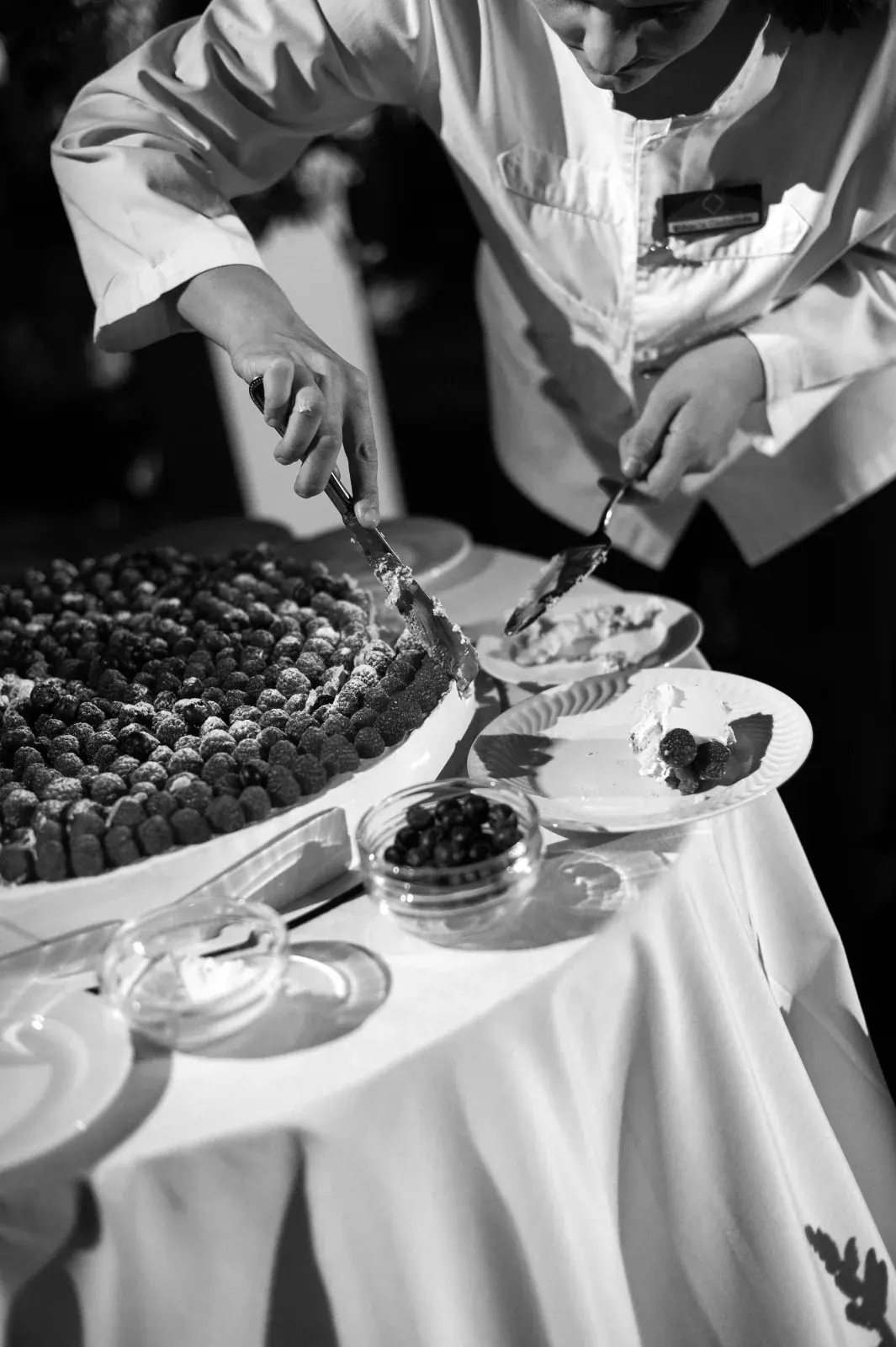 Chef slicing the berry-topped wedding cake at the table as service begins, in black and white