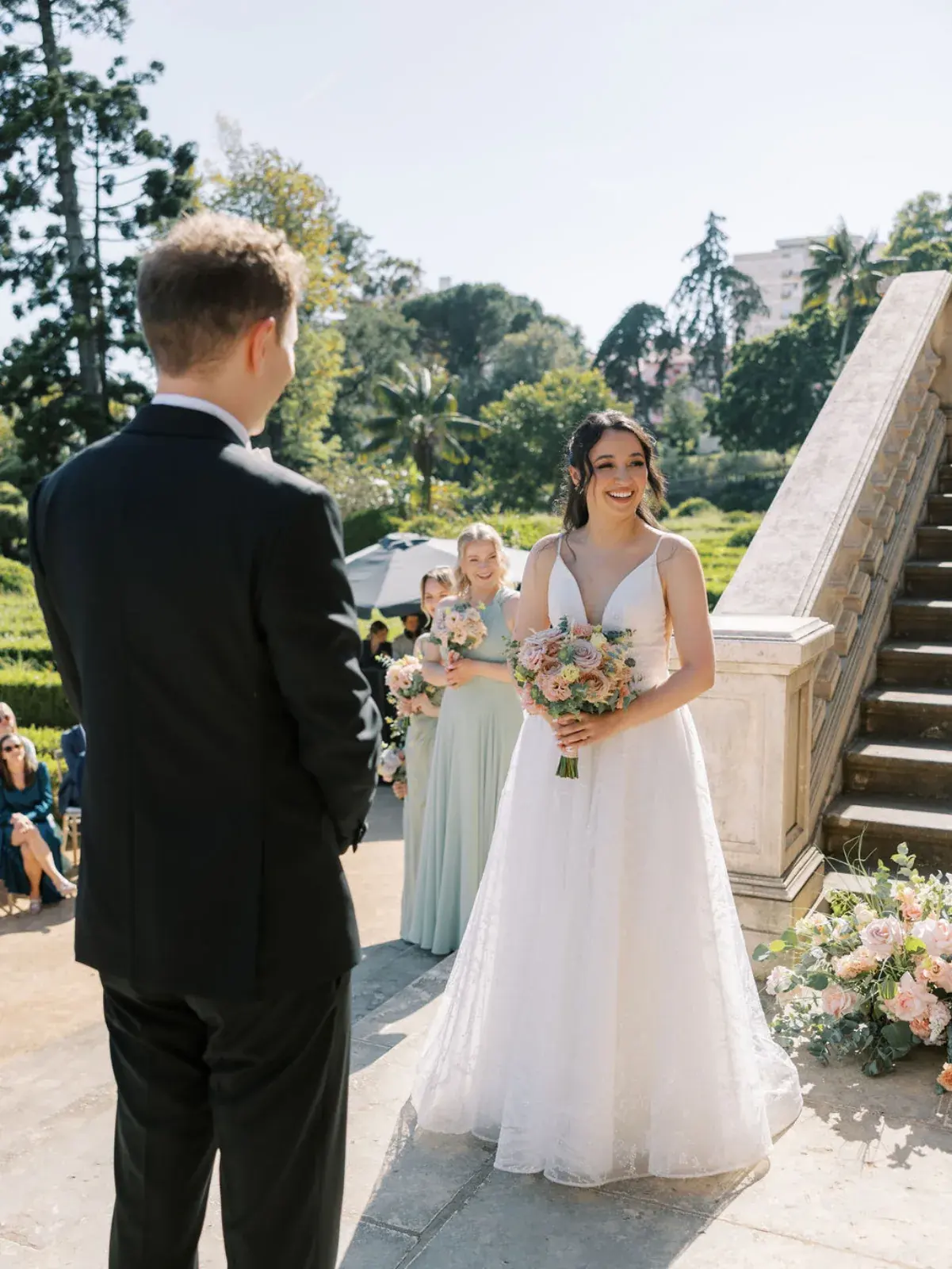 Groom turning to the bride at the foot of a stone garden staircase, her bouquet in blush tones