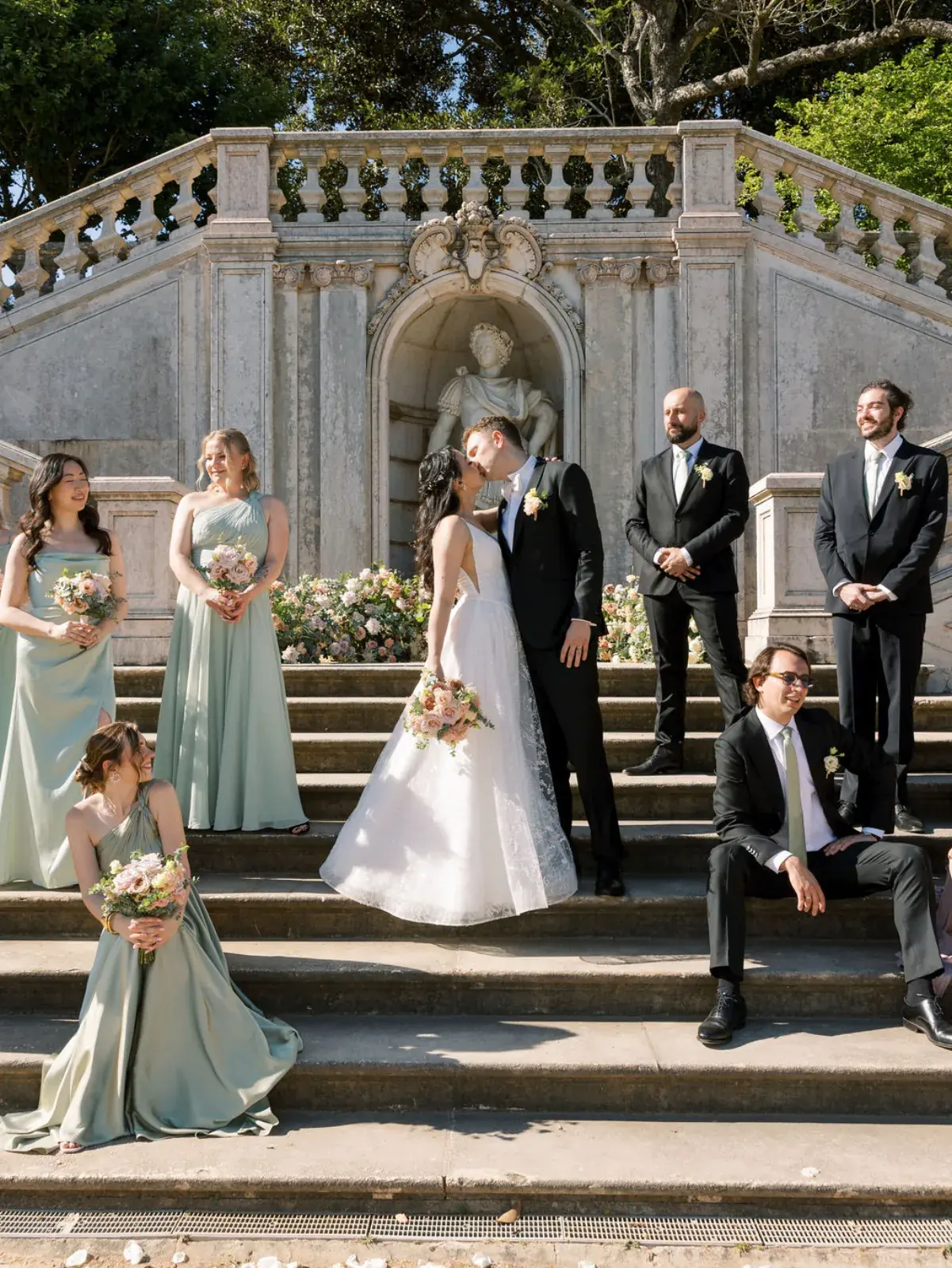 Wedding party in sage green flanking the couple's kiss on a baroque garden staircase