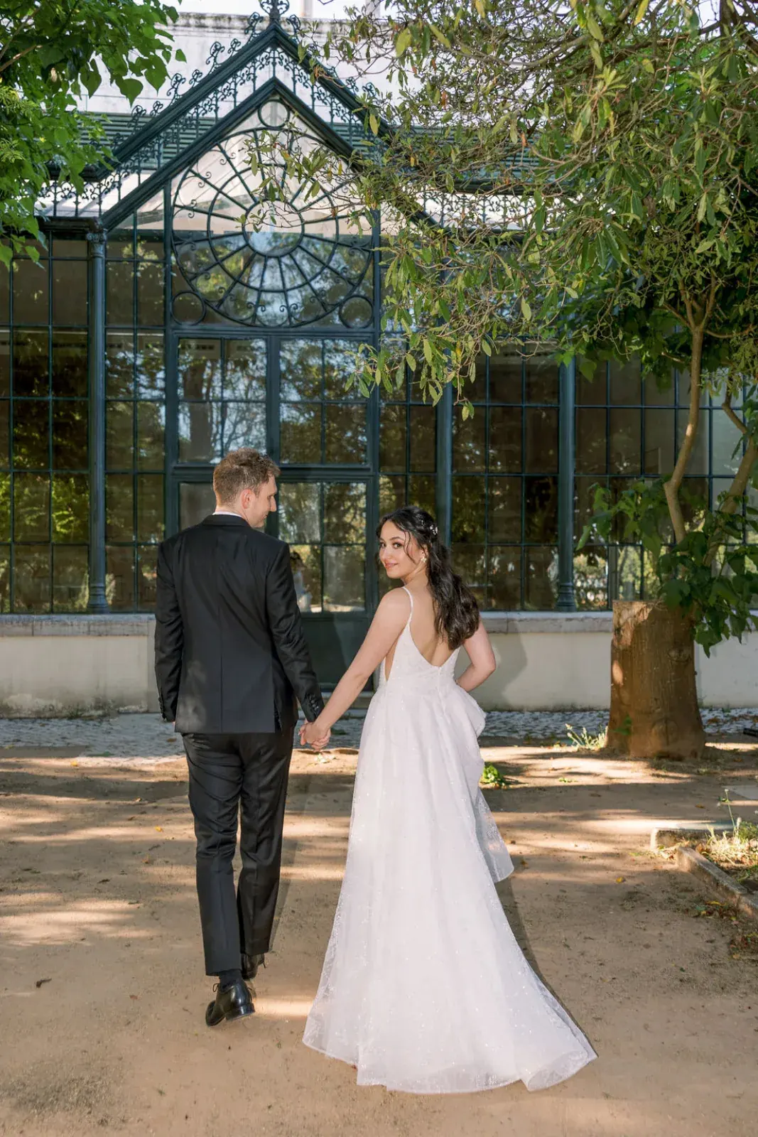 Couple walking toward a Victorian iron-and-glass greenhouse through the garden's afternoon shade
