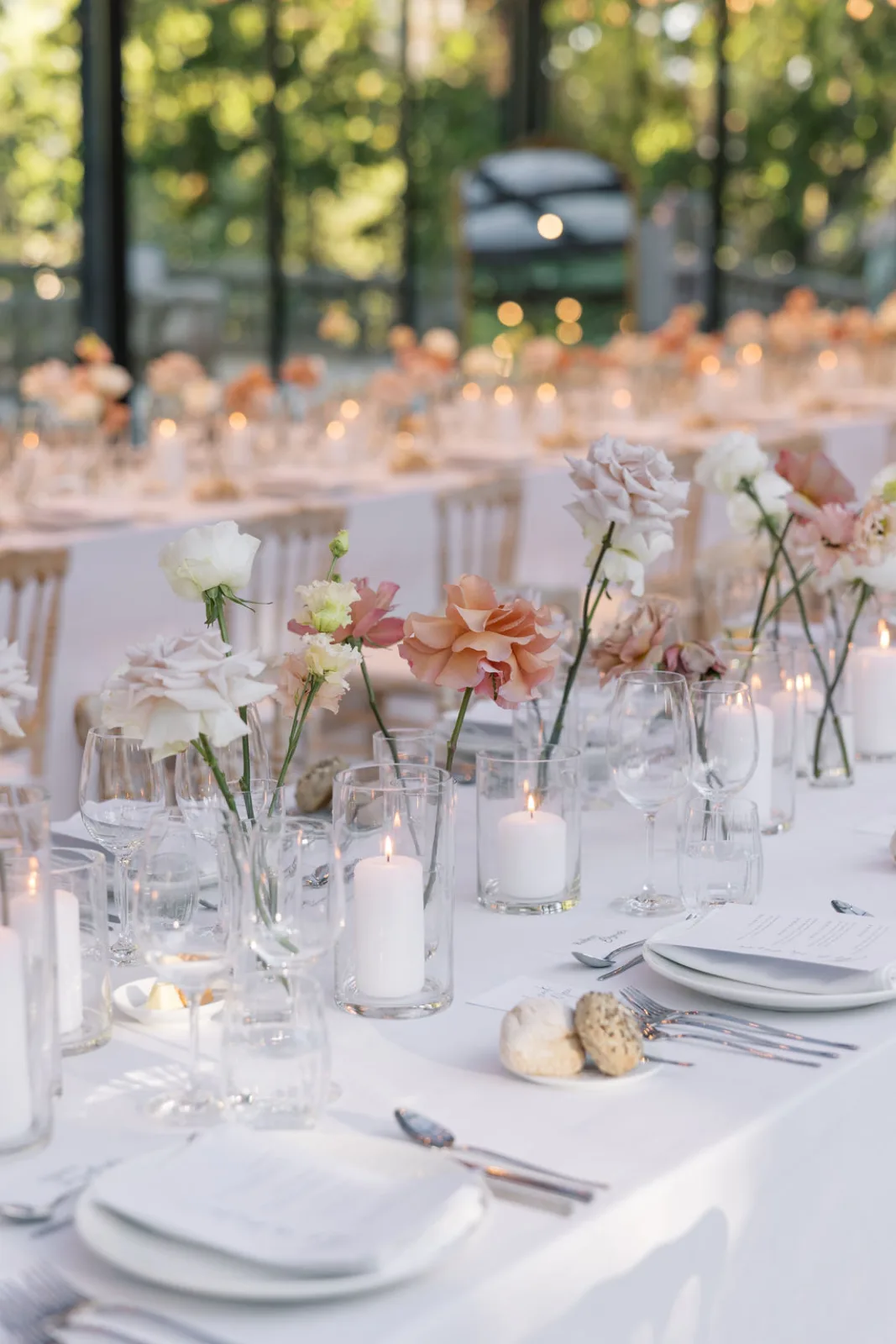 Reception tables inside the glasshouse, bud vases of toffee roses and pillar candles down the linen