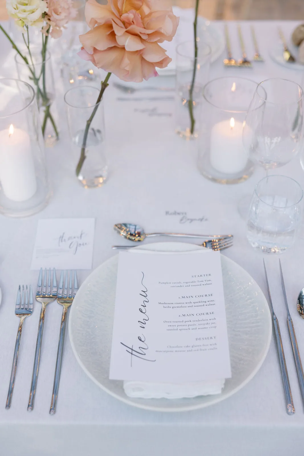 Script menu on a glass charger with gold cutlery, bud vases and candlelight along the table