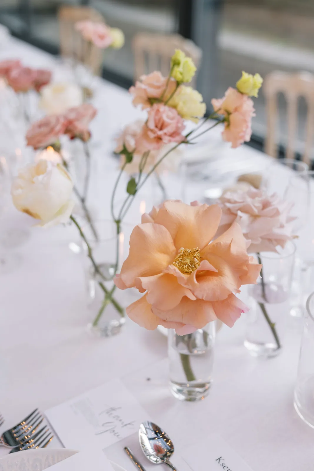 Bud vases of blush and peach roses along a white reception table, candles lit for the evening