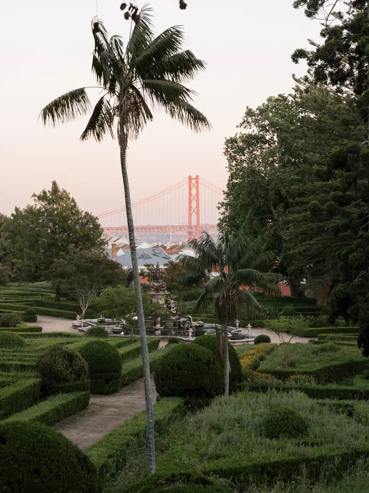 Dusk over box parterres and a garden fountain, the 25 de Abril bridge faint in the distance