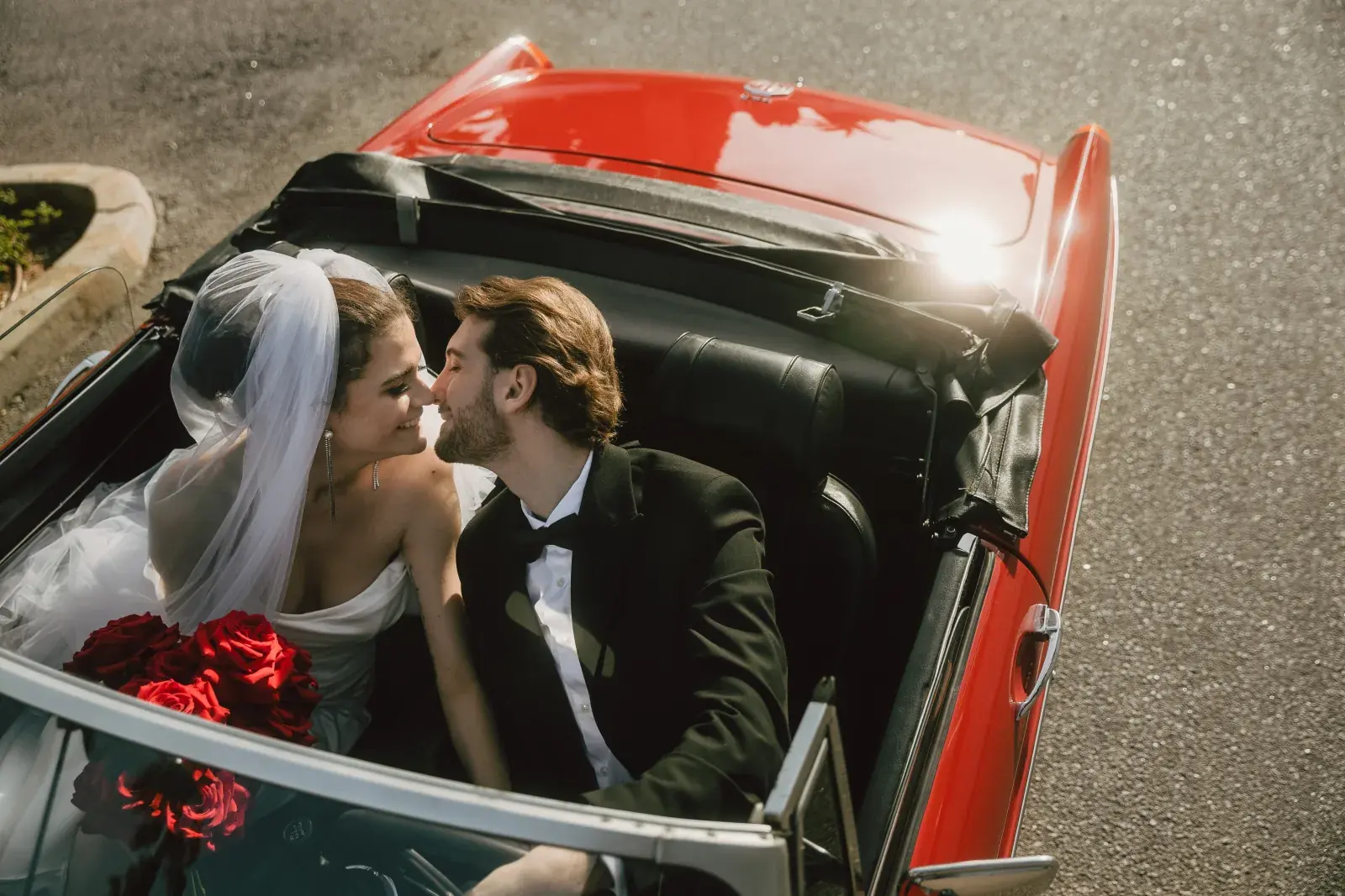 Bride and groom close together in the back of a red vintage convertible, red roses in hand