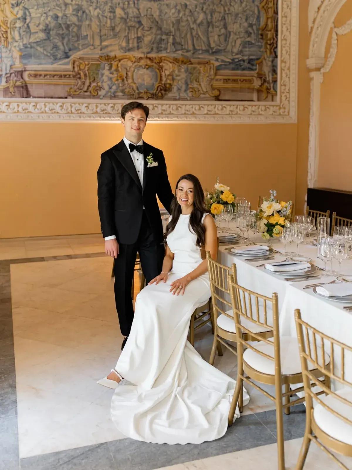 Couple beside a long table in an azulejo-panelled hall, yellow flowers down the runner