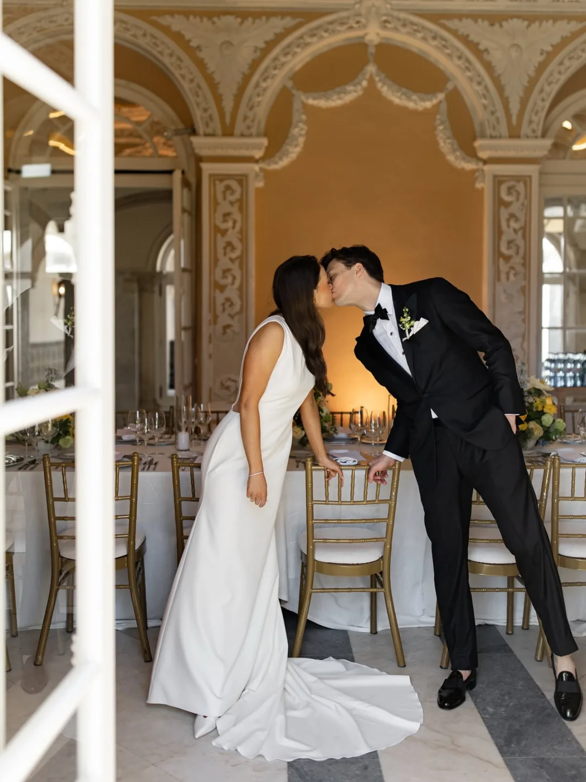 Bride and groom kissing across the banquet table in an arched hall of ornate stucco and warm ochre walls