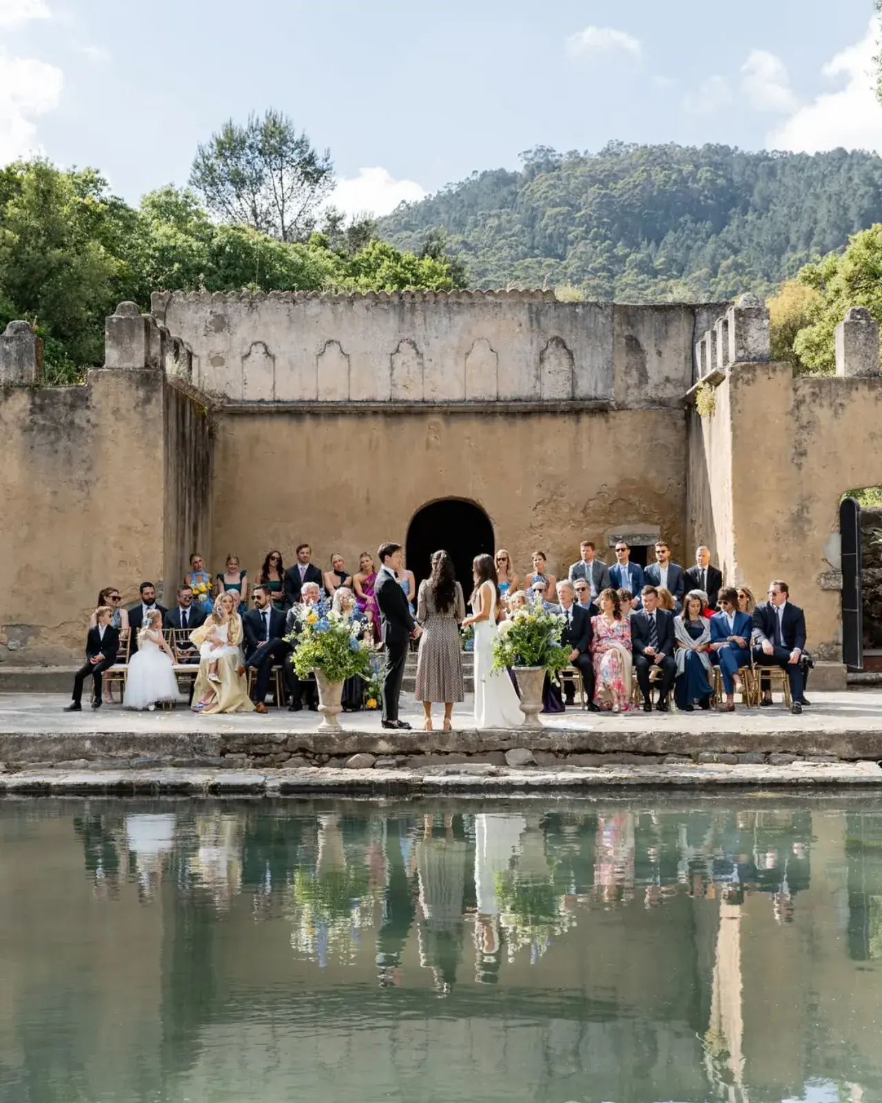 Ceremony on the ledge of an ancient walled water tank, guests and greenery mirrored in the pool
