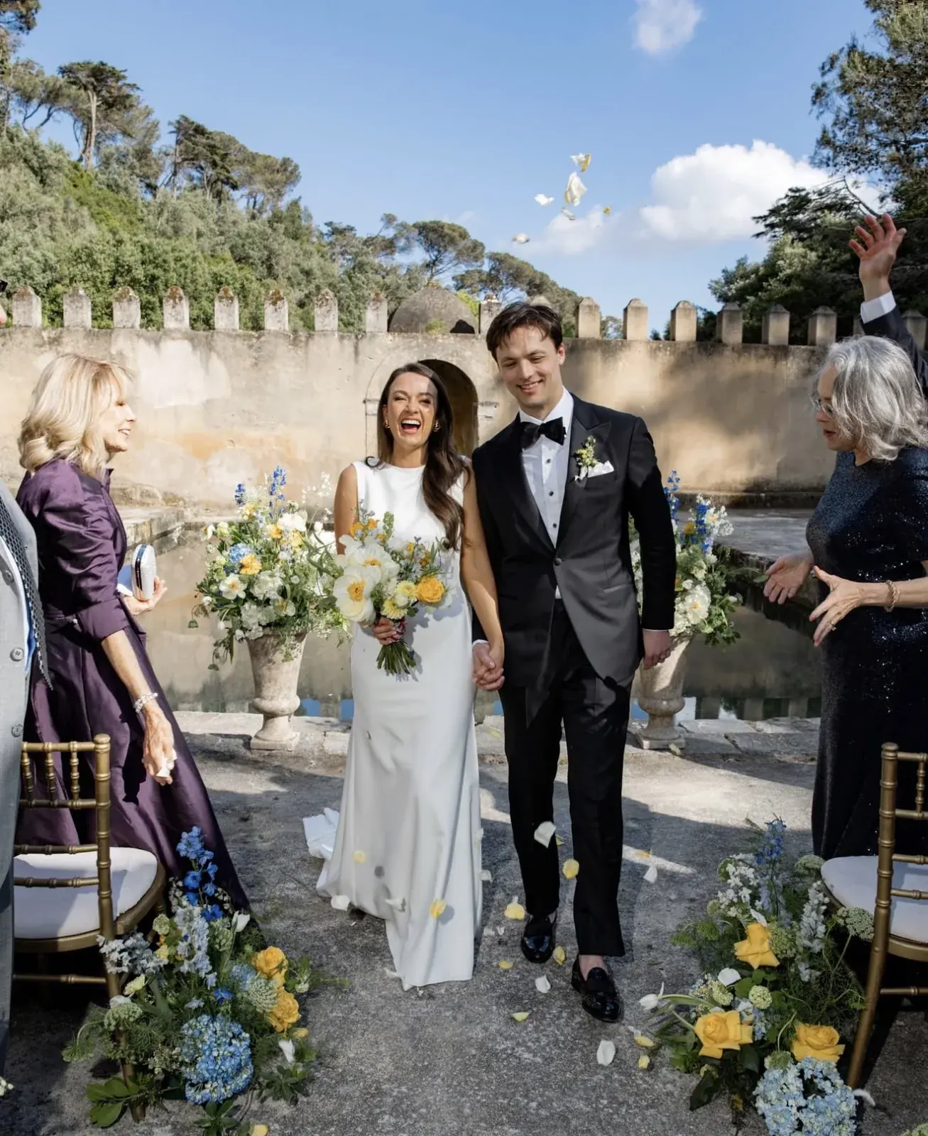 Newlyweds cheered along the aisle beneath castellated stone walls, petals in the air and gold chairs either side