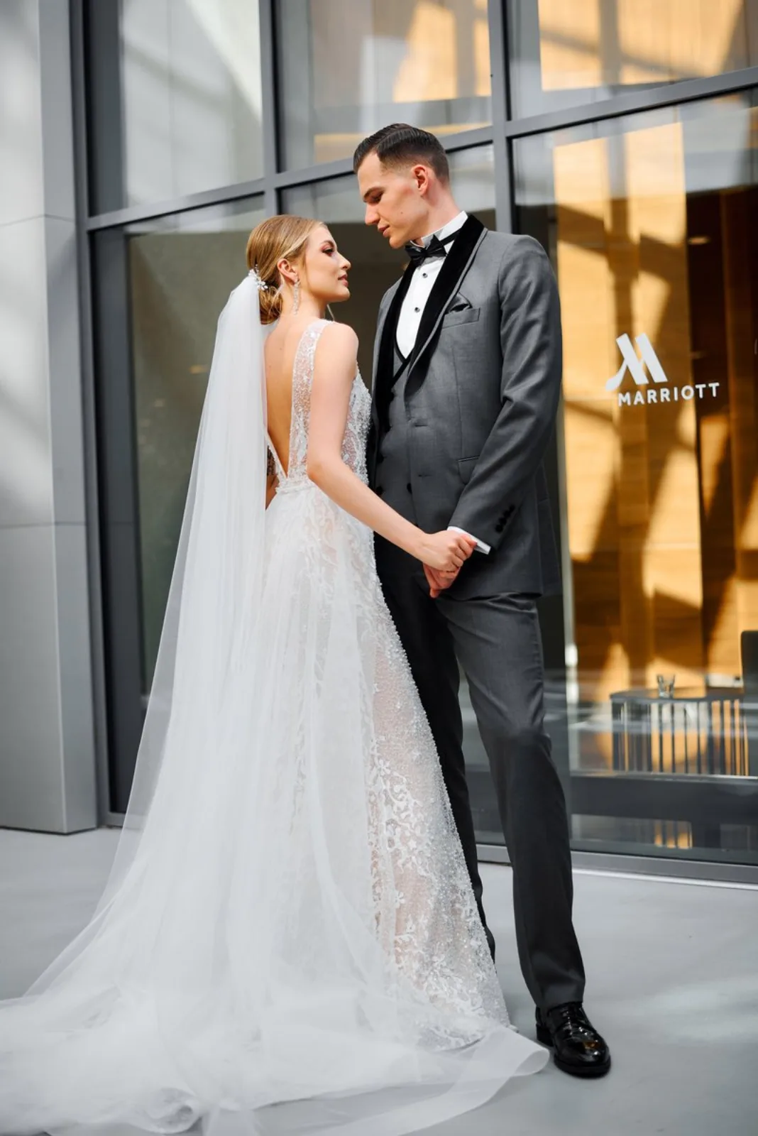 Bride and groom holding hands outside a contemporary glass-fronted hotel, veil catching the light