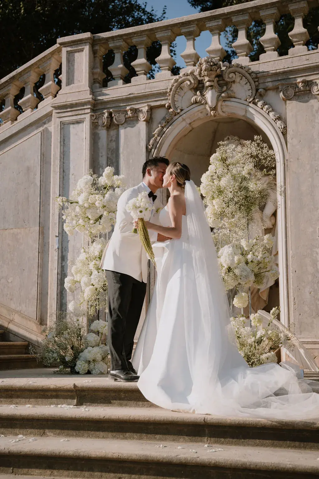 Couple kissing on palace steps under white hydrangea arch