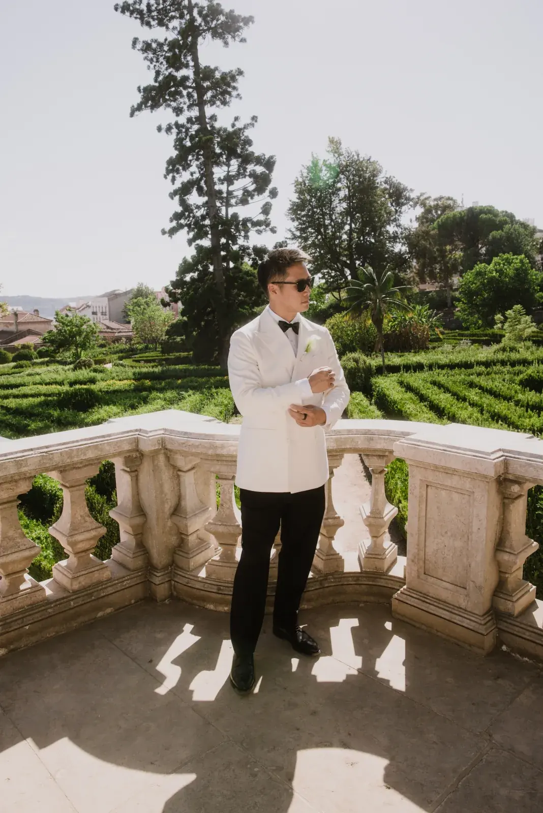 Groom in a white dinner jacket on a stone balcony overlooking geometric box hedging at midday