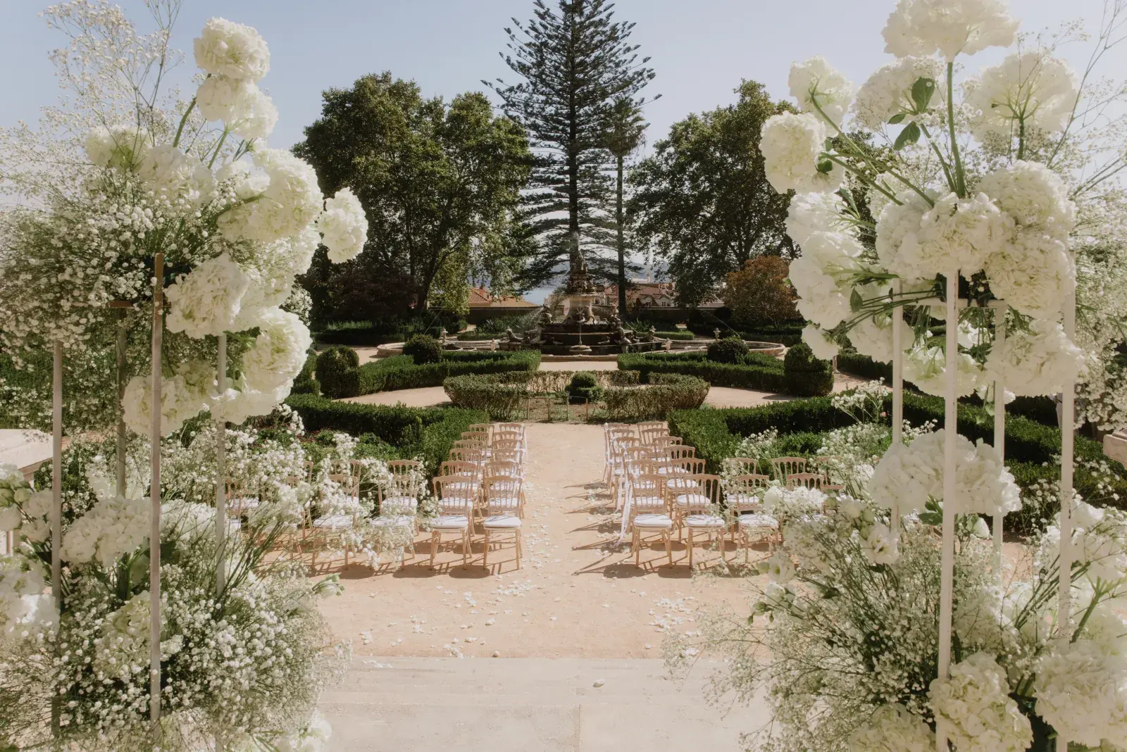 Ceremony encircling a fountain parterre, white floral columns framing the aisle beneath a great pine