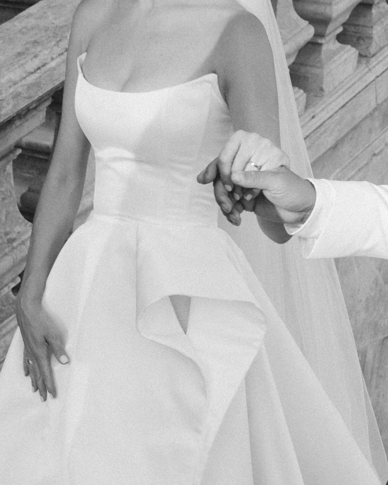 Structured folds of the bride's gown as the groom takes her hand by the balustrade, in black and white