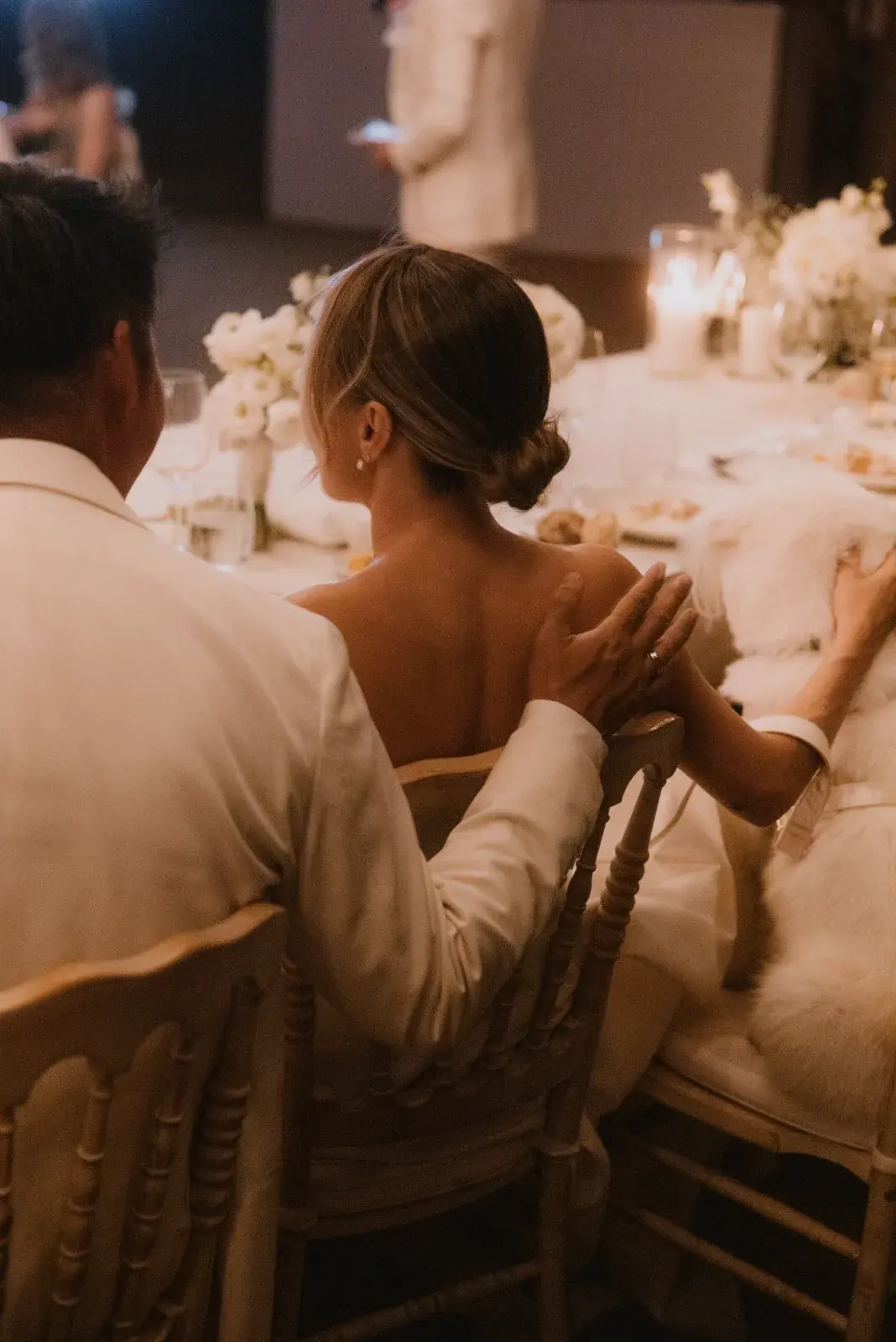 Newlyweds at a candlelit head table dressed with white flowers, the groom's hand on the bride's shoulder