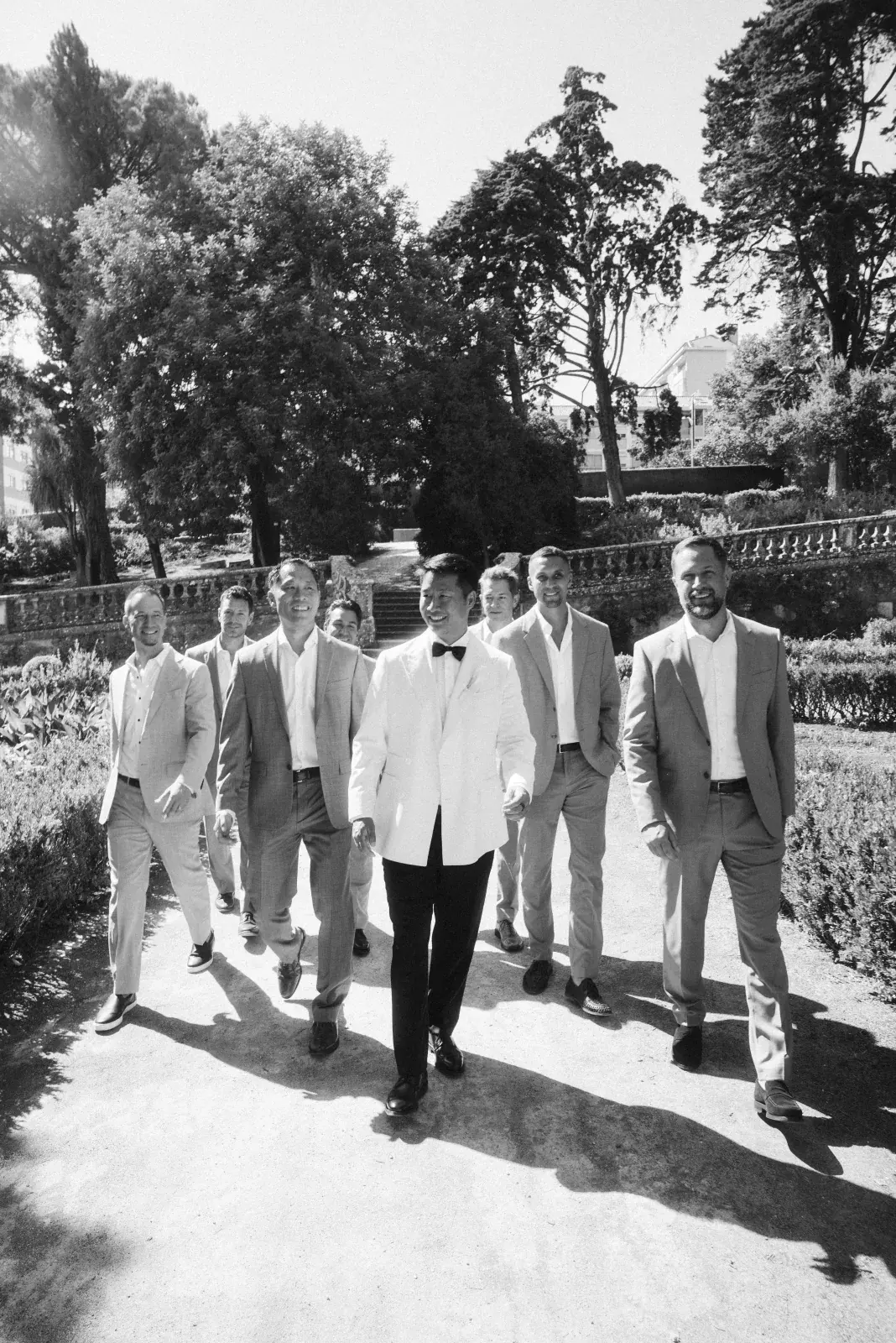 Groom in a white dinner jacket leading groomsmen through a formal garden in dappled midday light