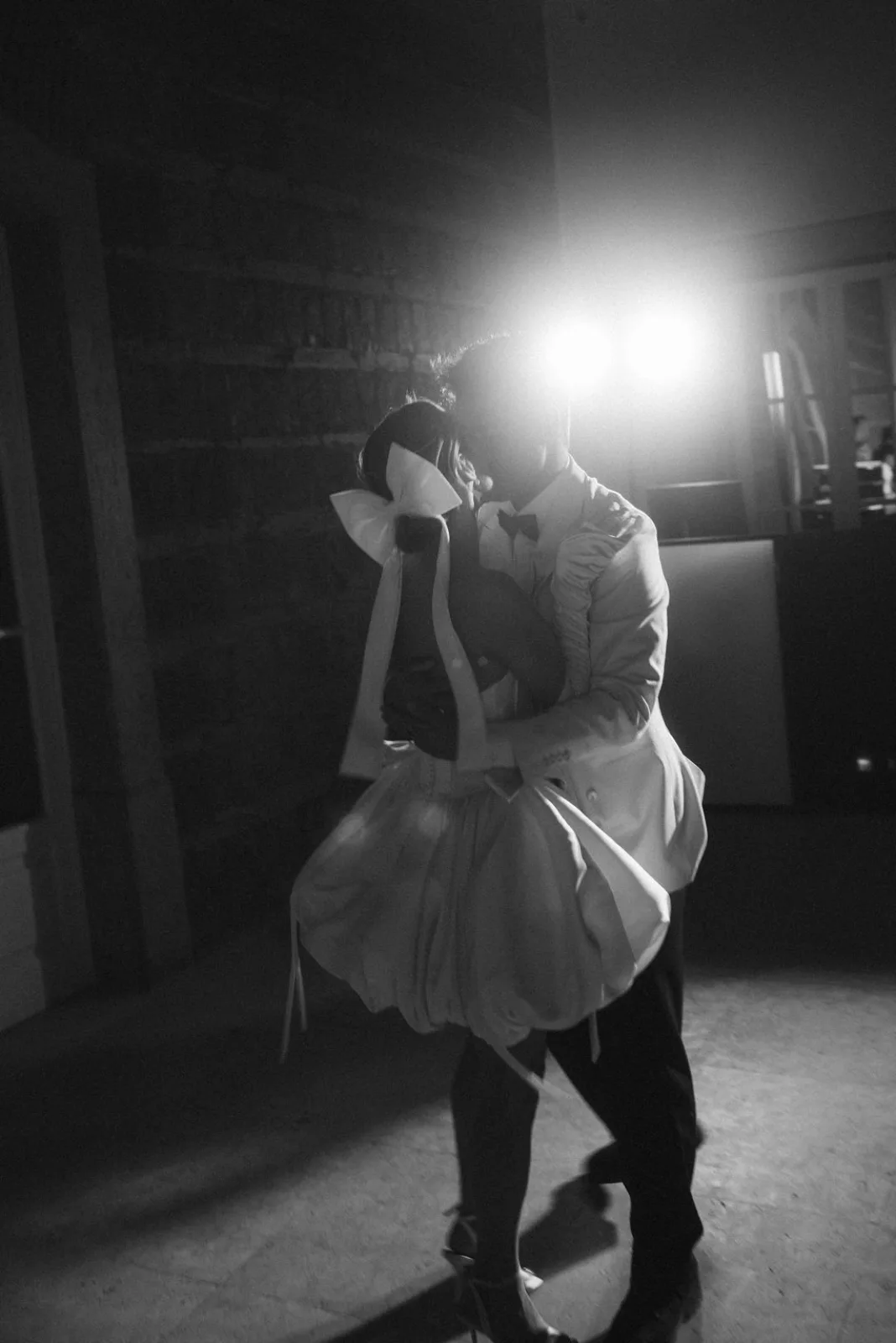 First dance in a darkened hall, backlight tracing the bow of the bride's gown, in black and white