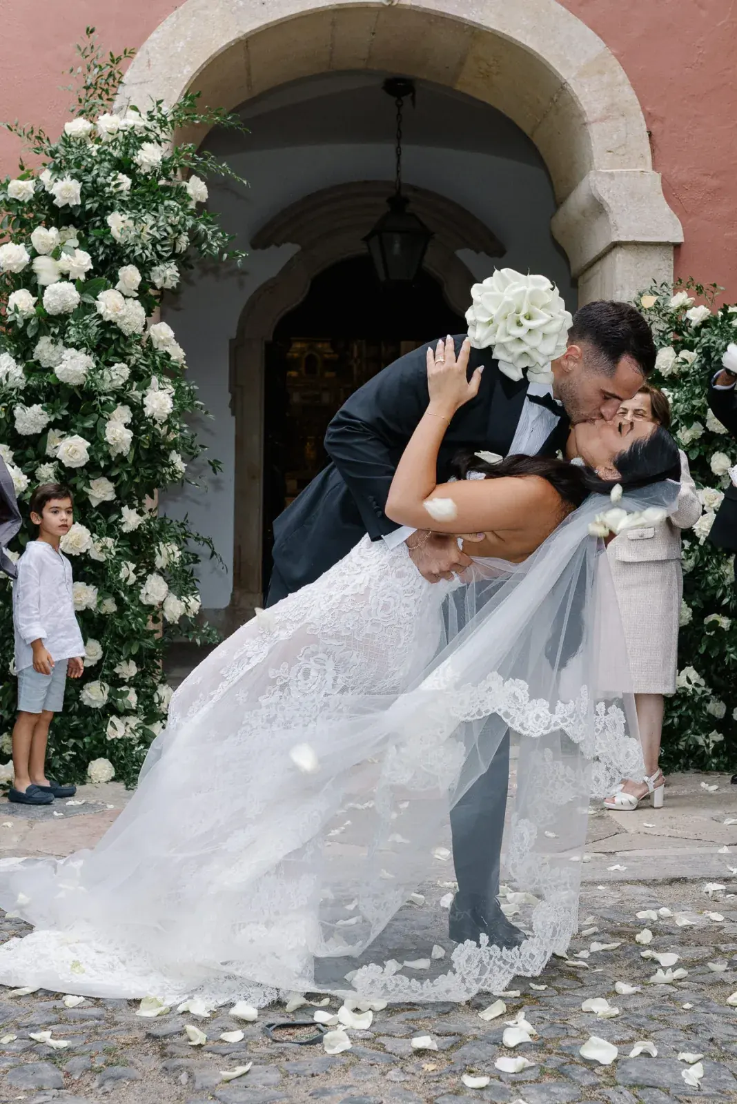 Groom dipping the bride in a kiss outside the chapel arch, white roses climbing the stone