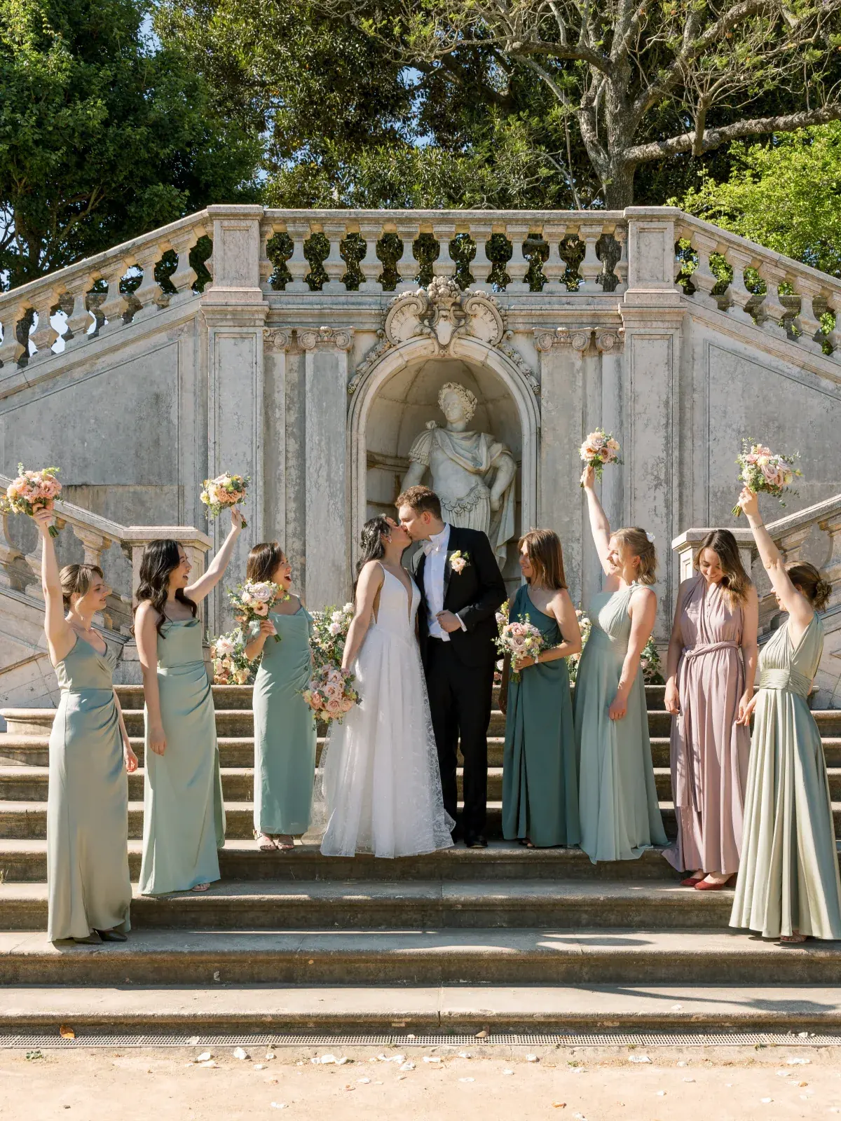 Bridesmaids in sage and rose raising bouquets around the couple on the palace garden steps