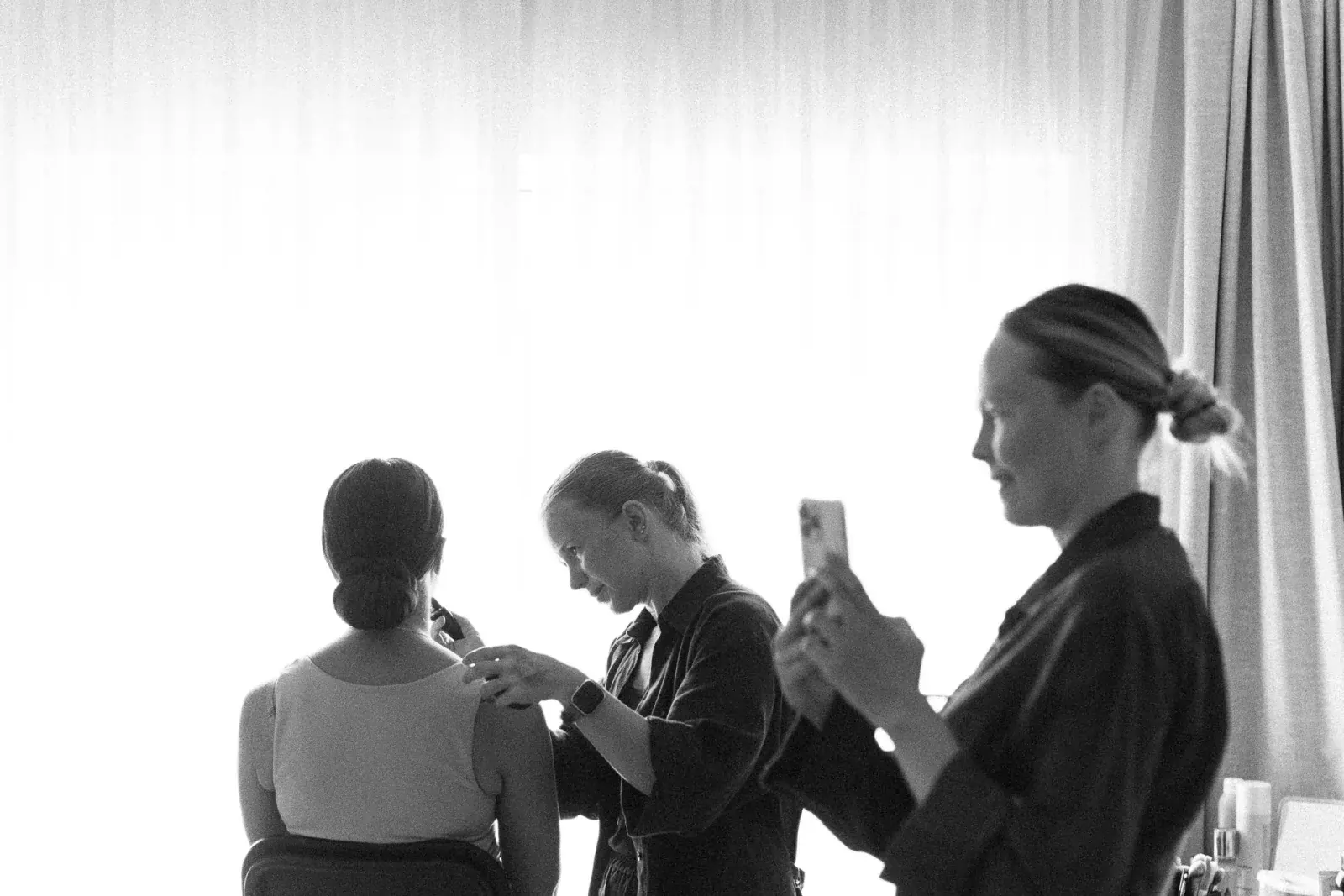 Bride at the window in soft daylight while stylists finish her hair, in black and white