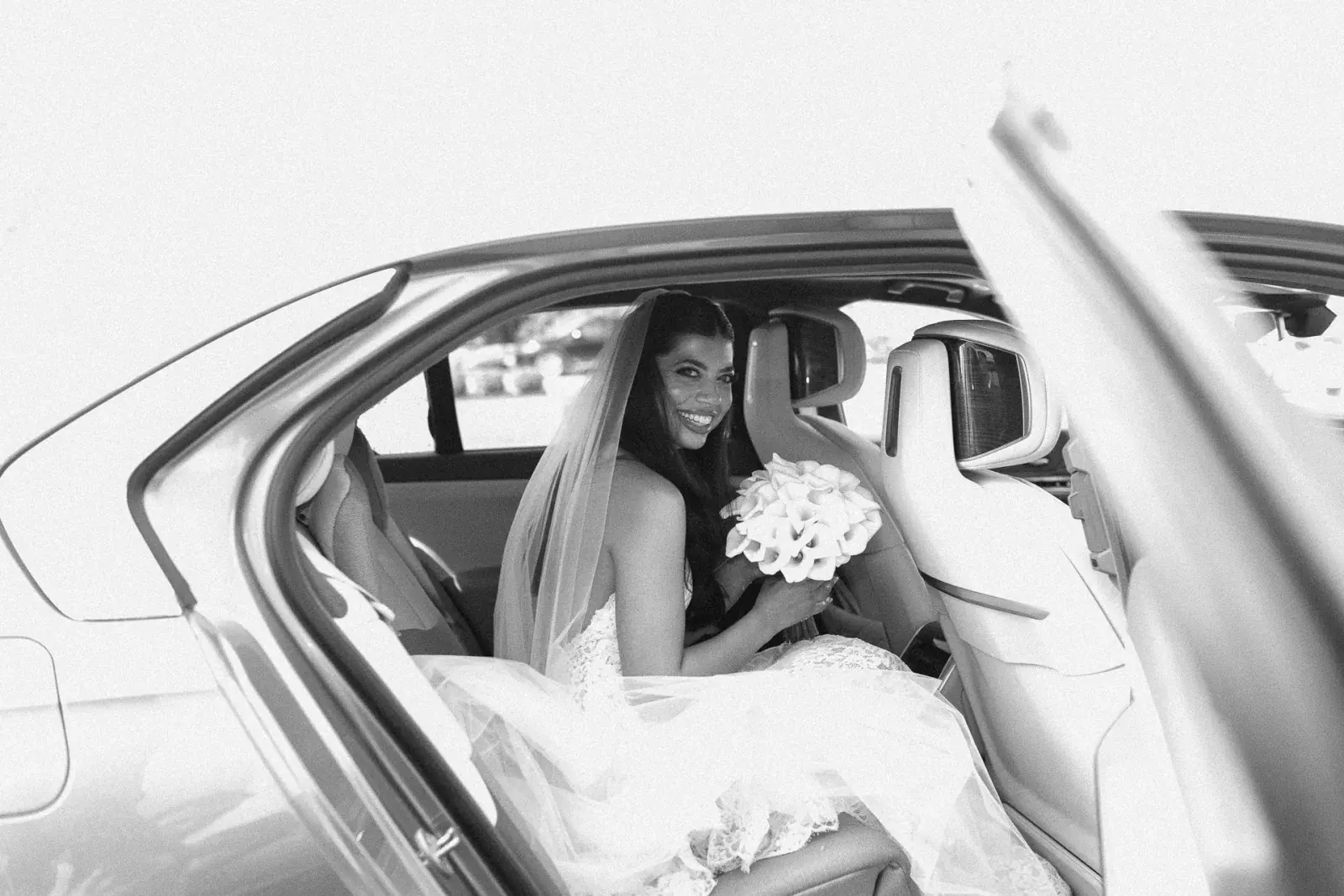 Bride in the wedding car with calla lilies, veil settled around her, in black and white