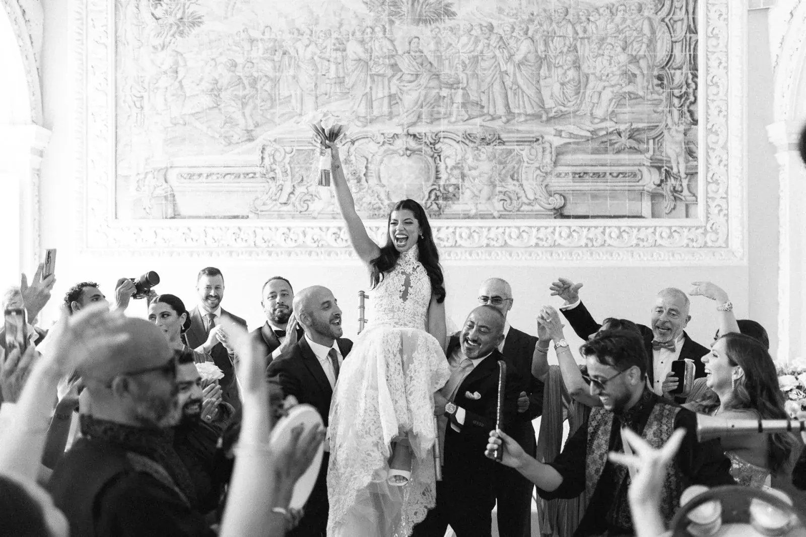 Bride lifted on shoulders with bouquet aloft, guests dancing beneath a monumental azulejo wall