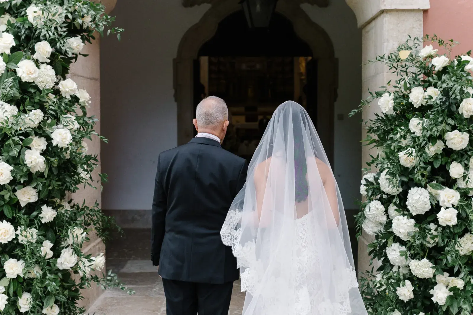 Father walking the bride through a stone chapel doorway framed by columns of white roses