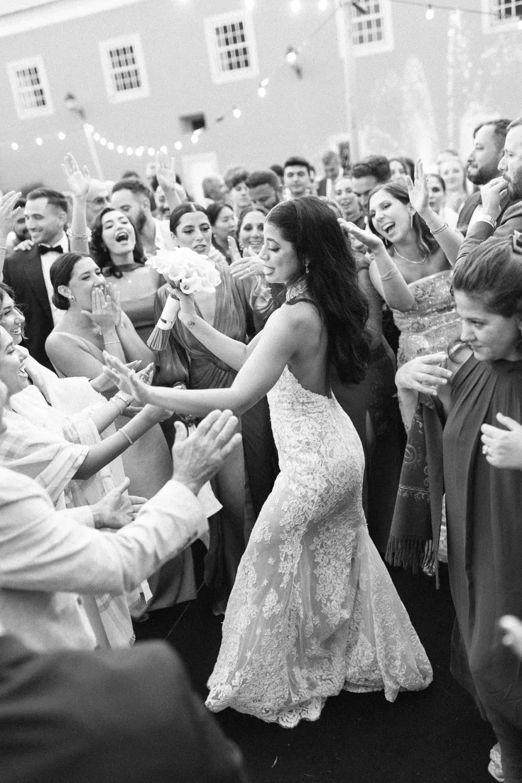 Bride in lace dancing at the centre of clapping guests beneath festoon lights, in black and white
