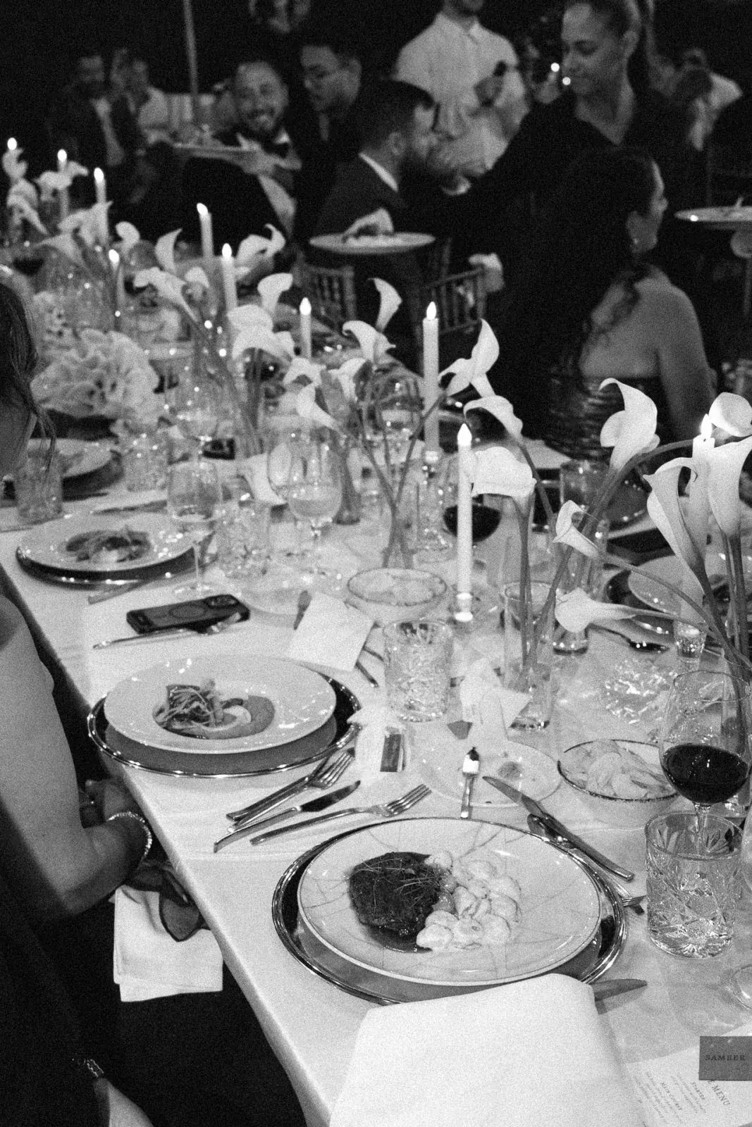 Calla lilies and taper candles down a night-time dinner table, guests deep in conversation