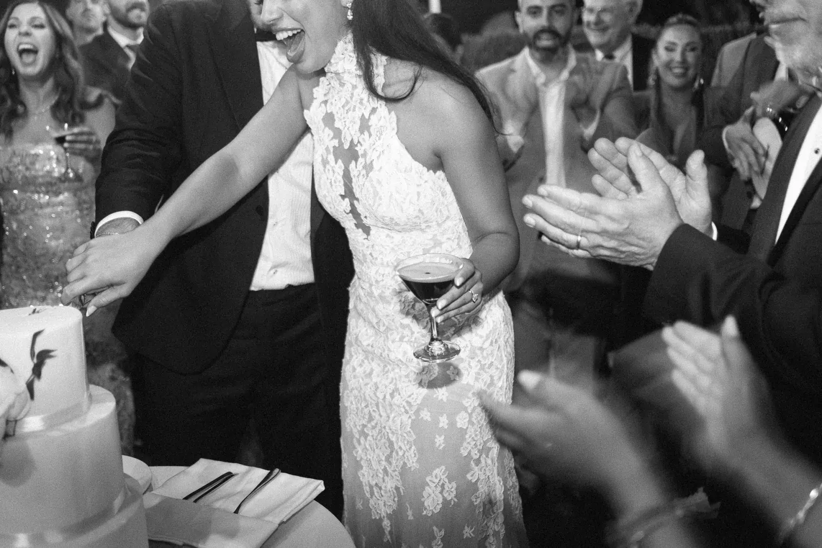 Bride laughing over an espresso martini as the cake is cut, guests applauding, in black and white