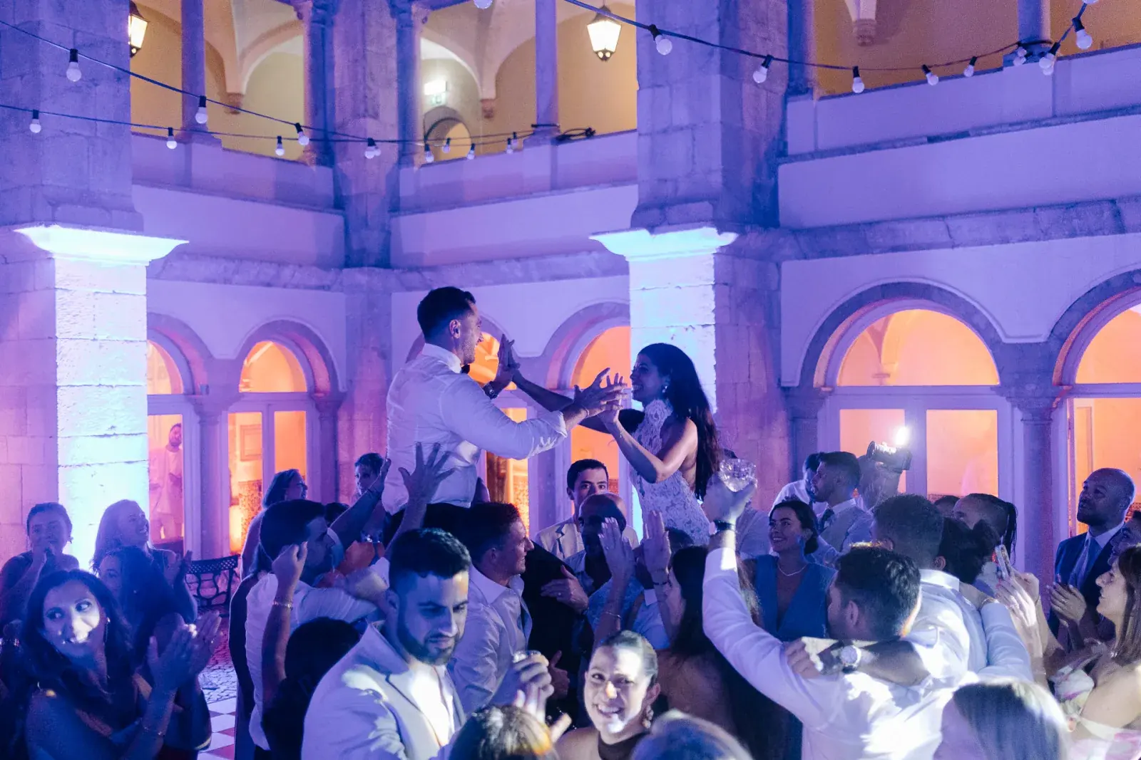 Guests dancing in an arcaded palace courtyard under festoon lights and violet evening lighting